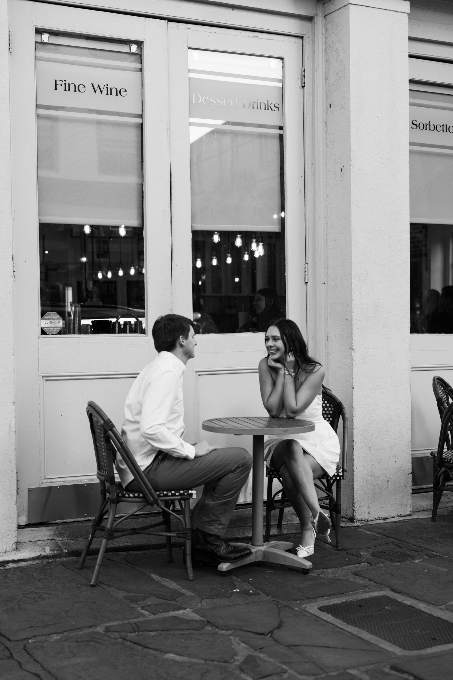 Black and white photo of a couple sitting at an outdoor cafe table in front of a fine wine shop.