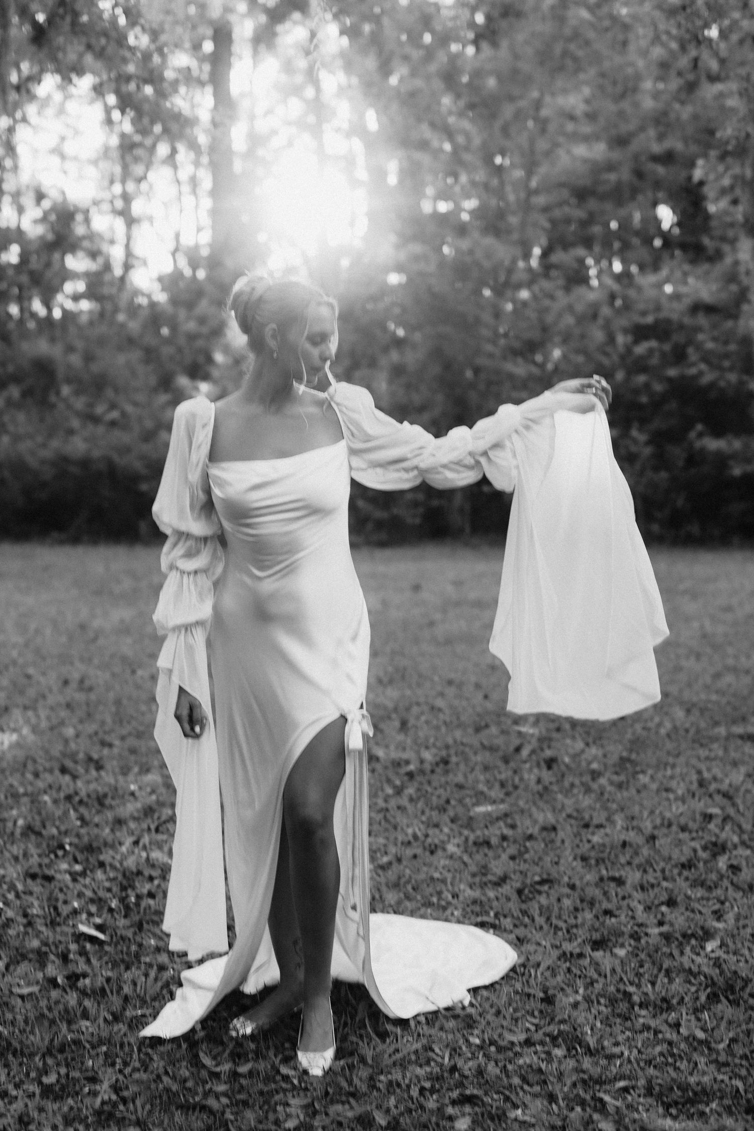 Black and white photo of a bride in a flowing white wedding dress holding her veil in sunlit outdoor garden.