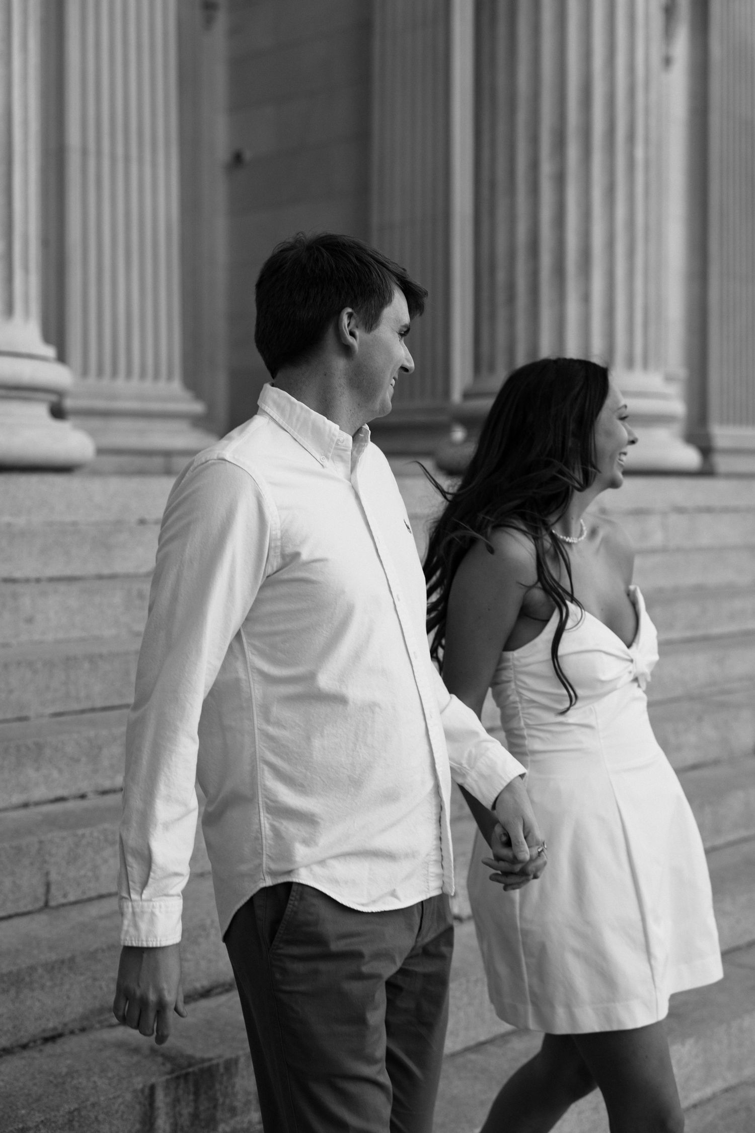 Black and white photo of a couple holding hands on steps near classical columns.