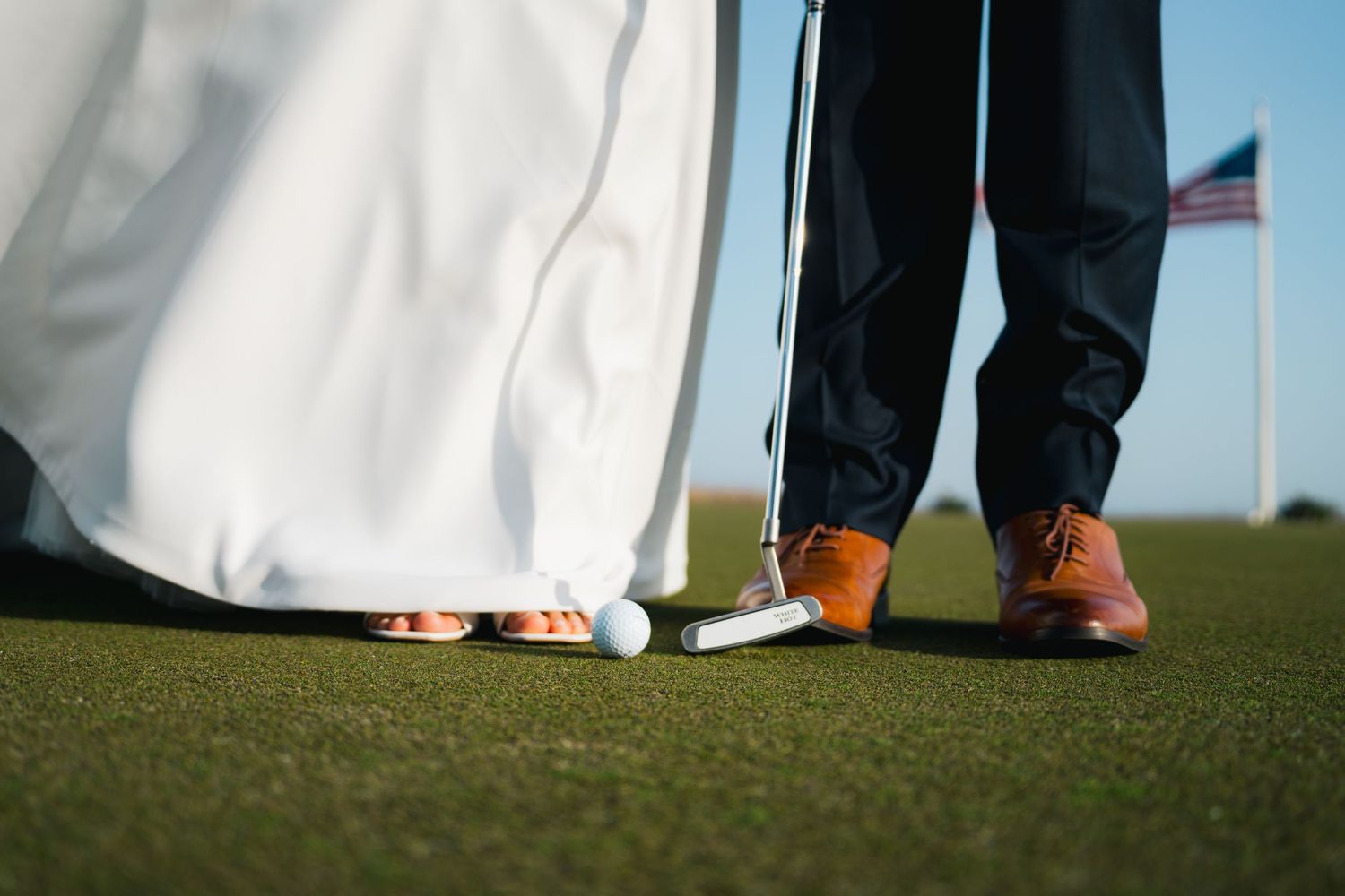 Close-up of bride and groom's feet on golf course green with golf ball and American flag in background.