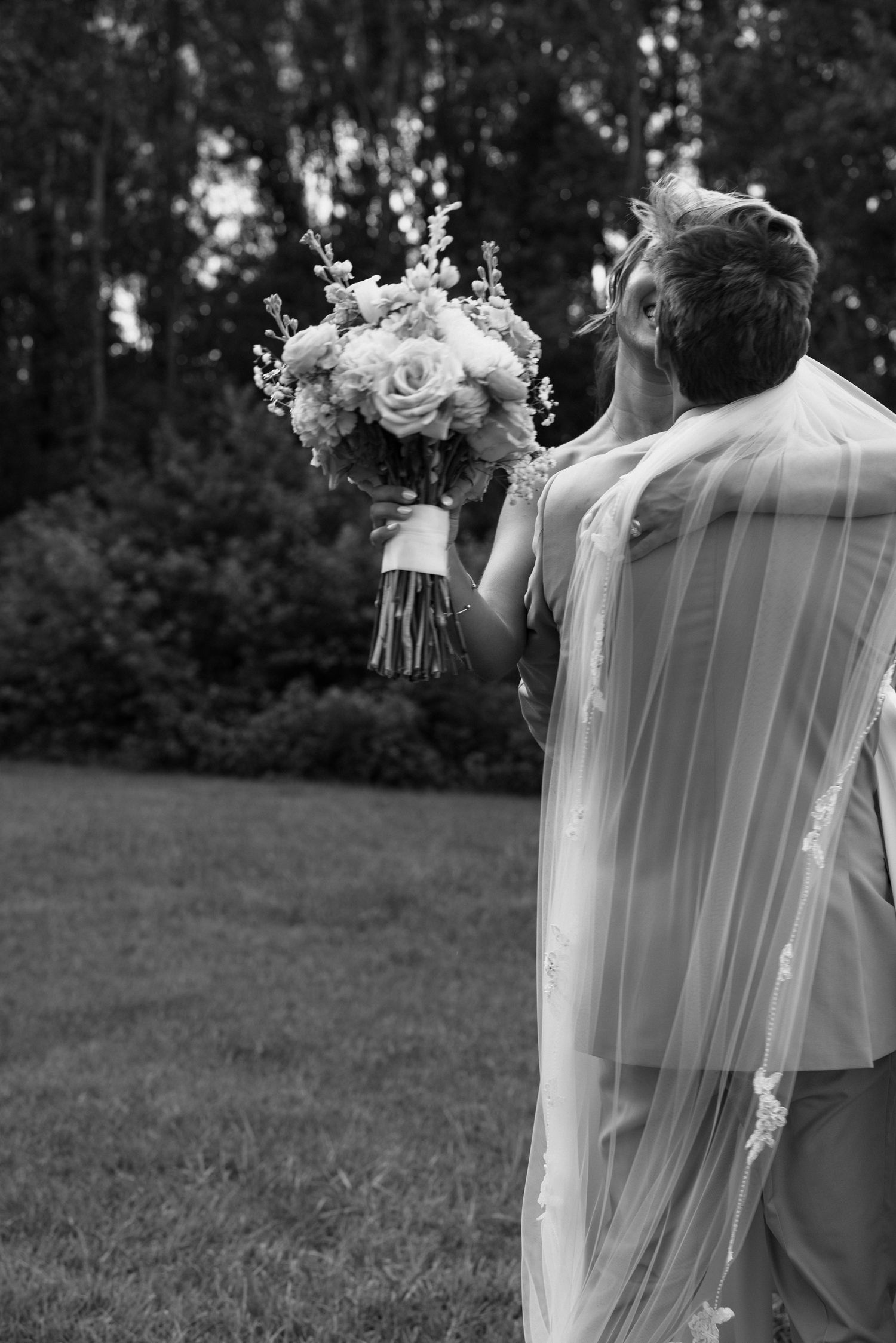 Black and white photo of a bride and groom embracing outdoors, bride holding a floral bouquet with veil flowing.