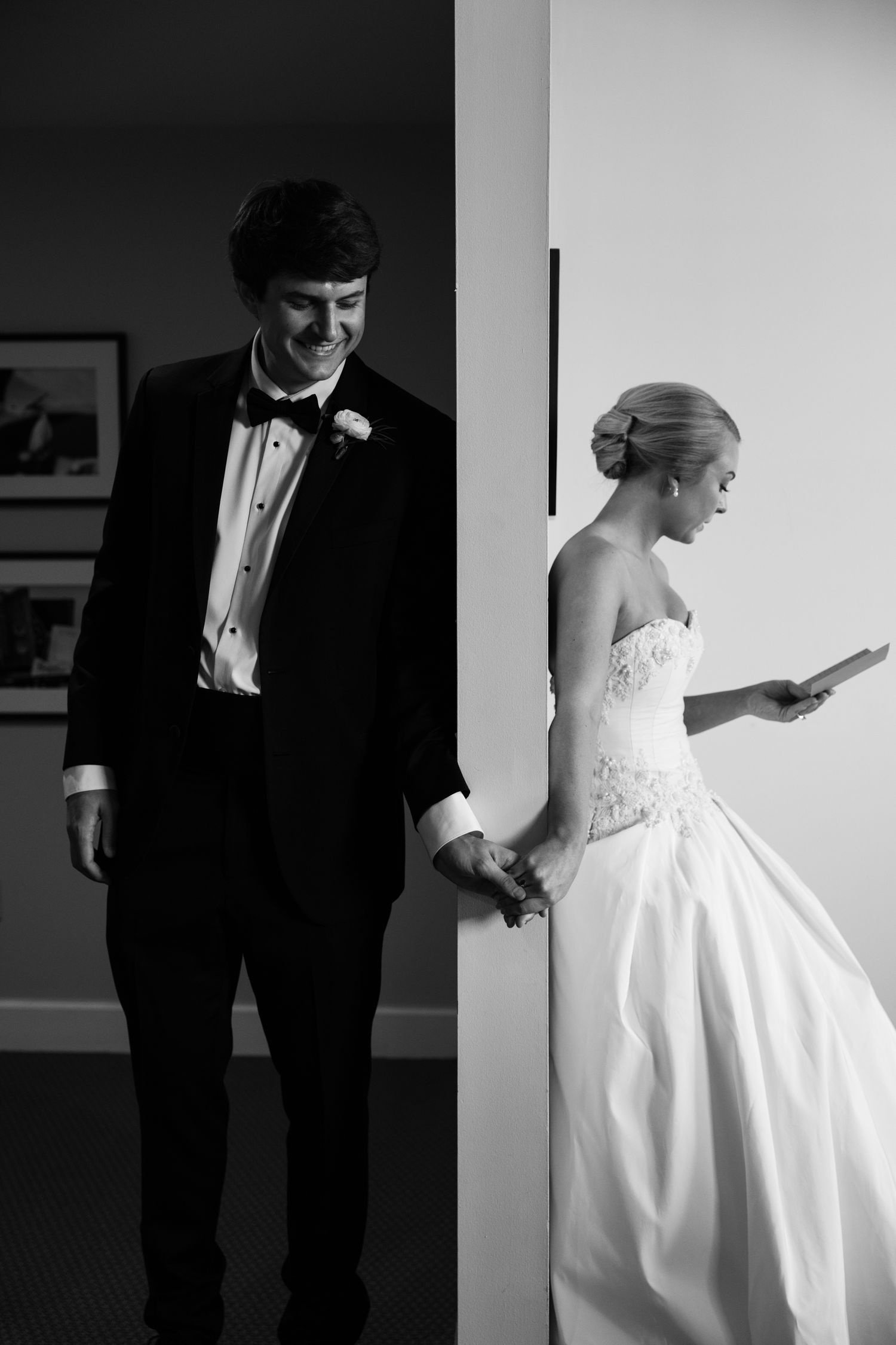Black and white photo of bride and groom holding hands around a wall on their wedding day.