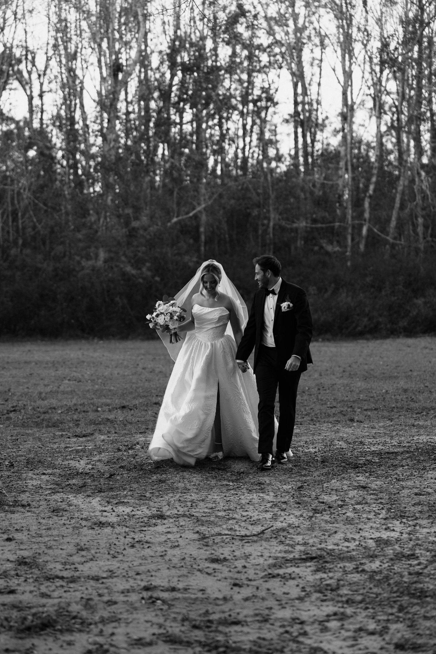 Black and white photo of a bride and groom walking hand in hand across a field with trees in the background.