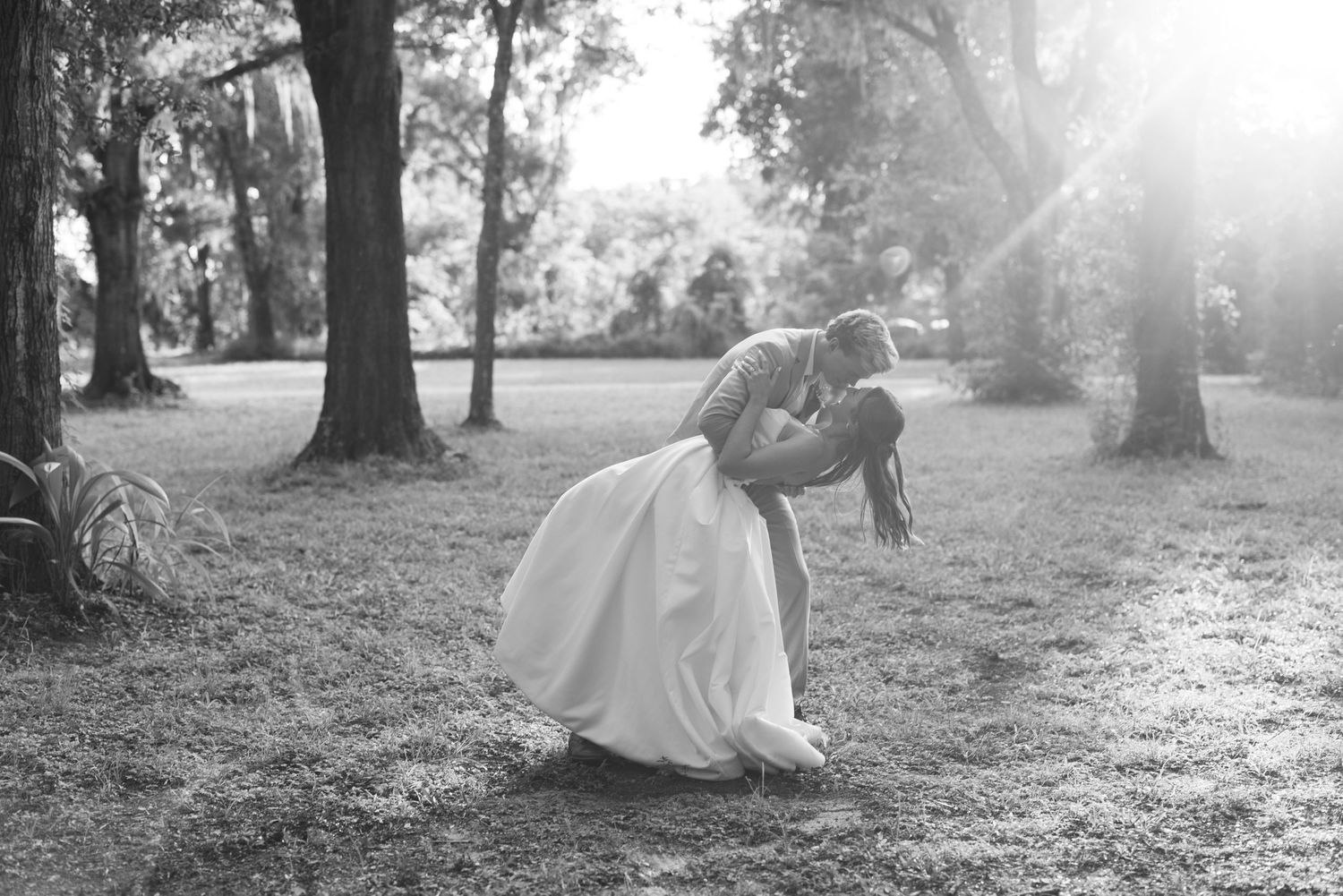 Black and white romantic wedding photo of couple sharing a dip kiss among sunlit trees in a garden.