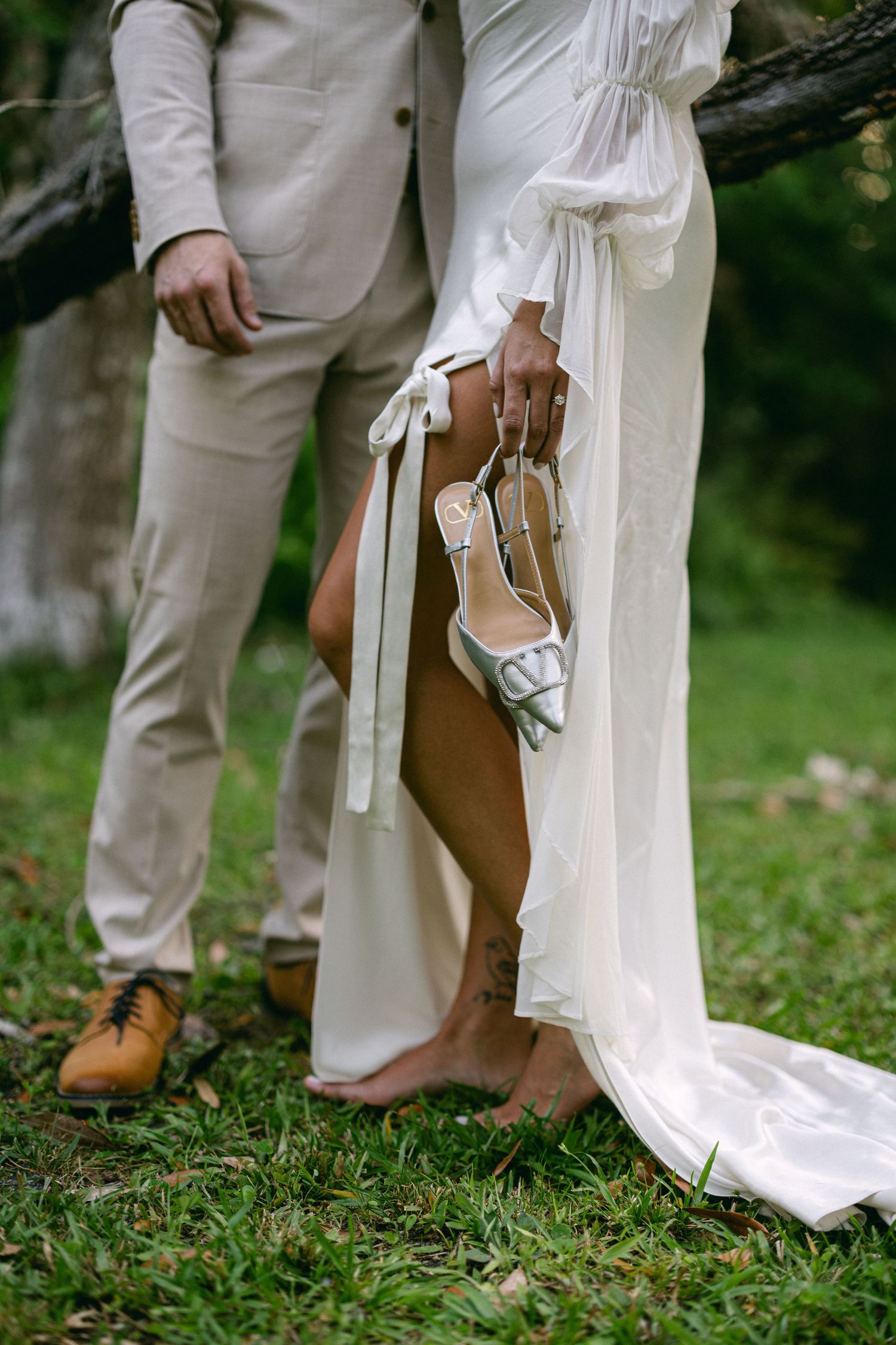 Barefoot bride holding white heels beside groom on grass, showcasing elegant wedding attire details.
