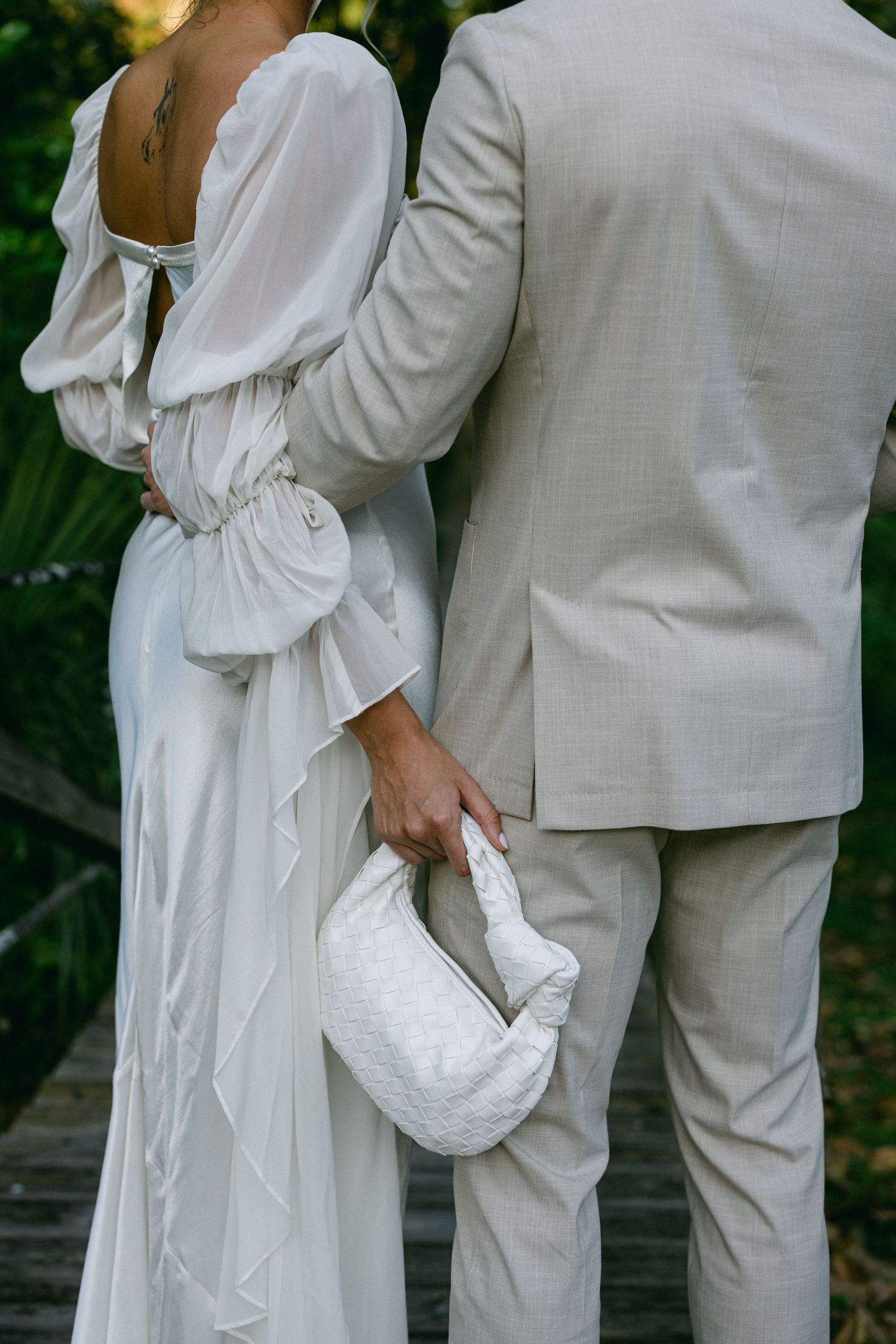 Close-up of bride in backless white gown holding woven bag beside groom in beige suit outdoors.