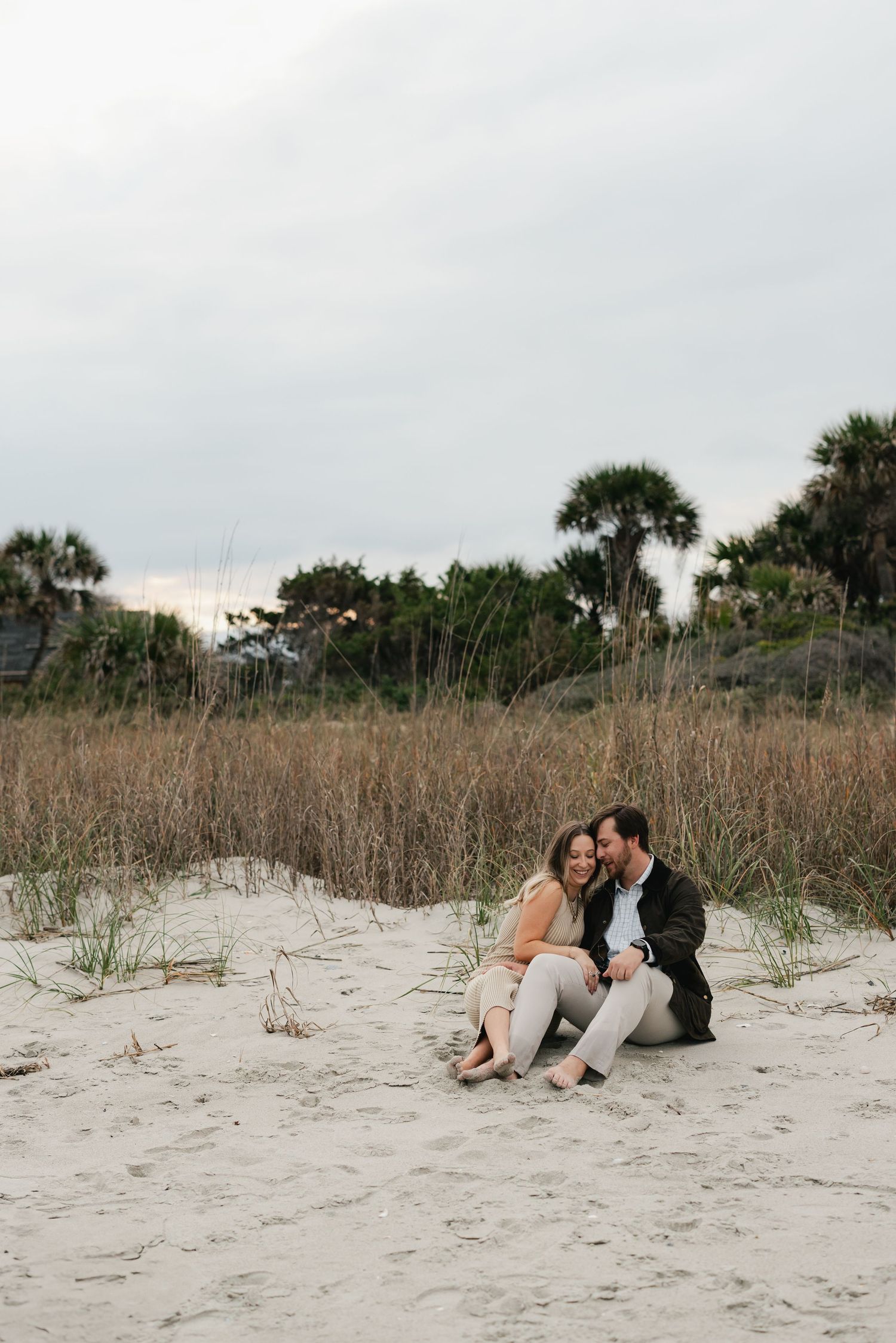 Couple sitting together on sandy beach dunes with palm trees and sea grass in background.