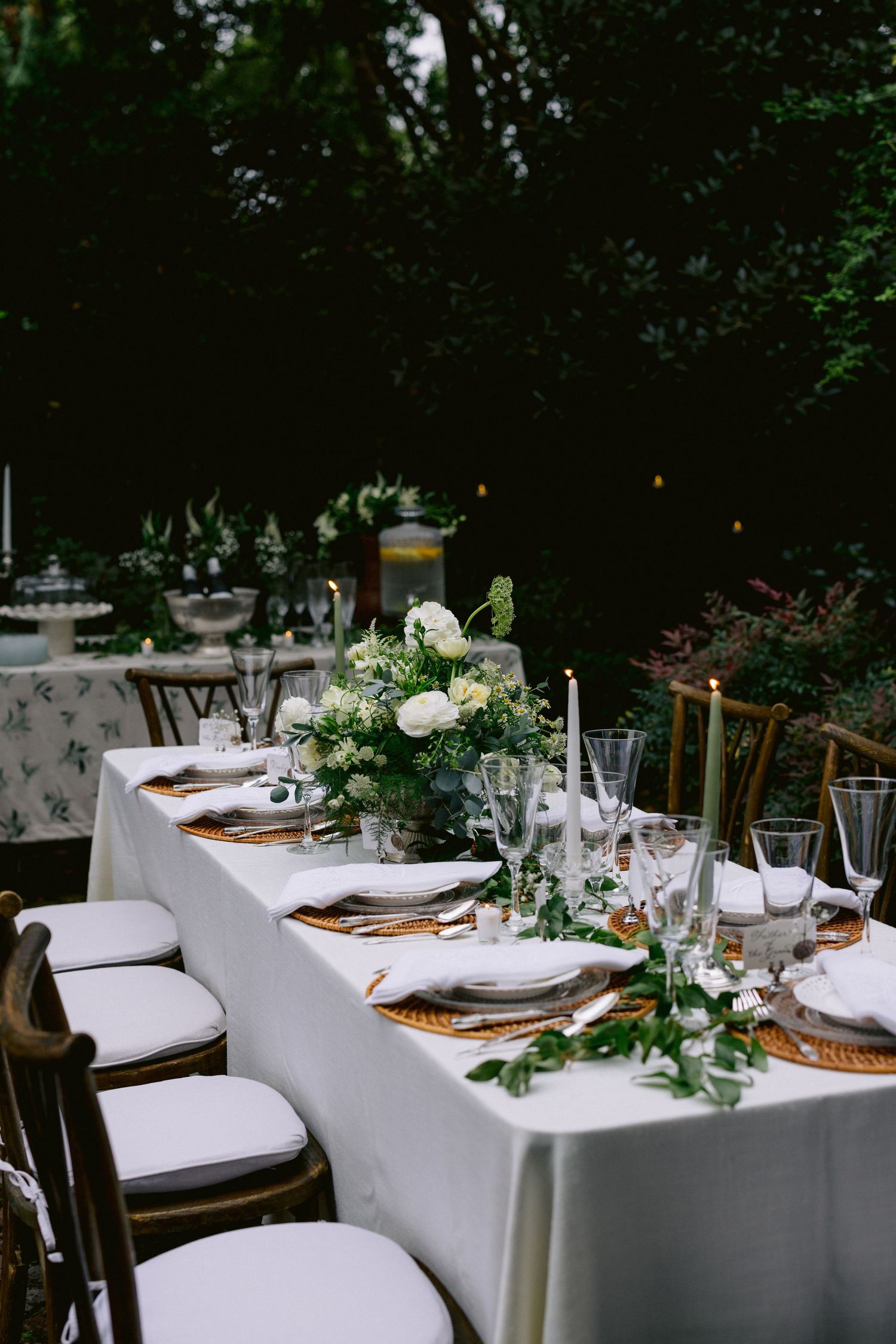 Romantic outdoor wedding table with white roses, greenery runner, taper candles, crystal glasses, and wicker chargers.