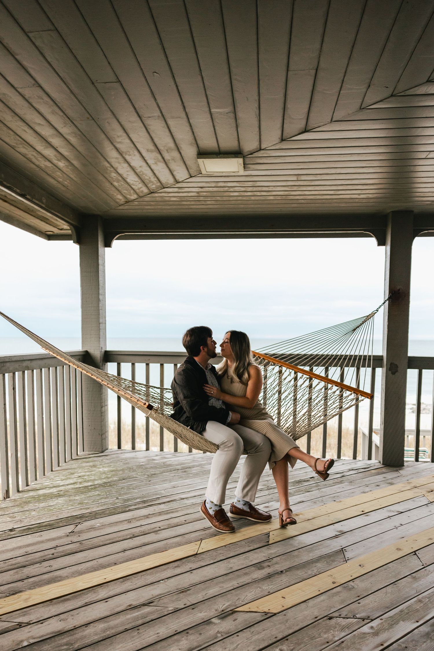 Couple sharing a romantic kiss on a hammock in a beachside wooden gazebo with ocean views.