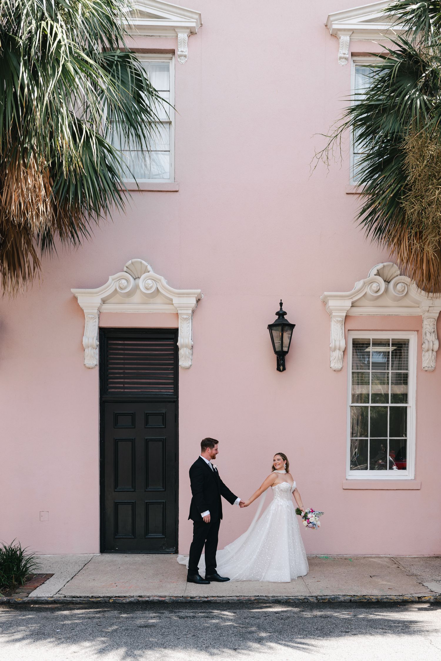 Bride and groom hold hands in front of a pink building with black door and palm trees in Charleston, SC.