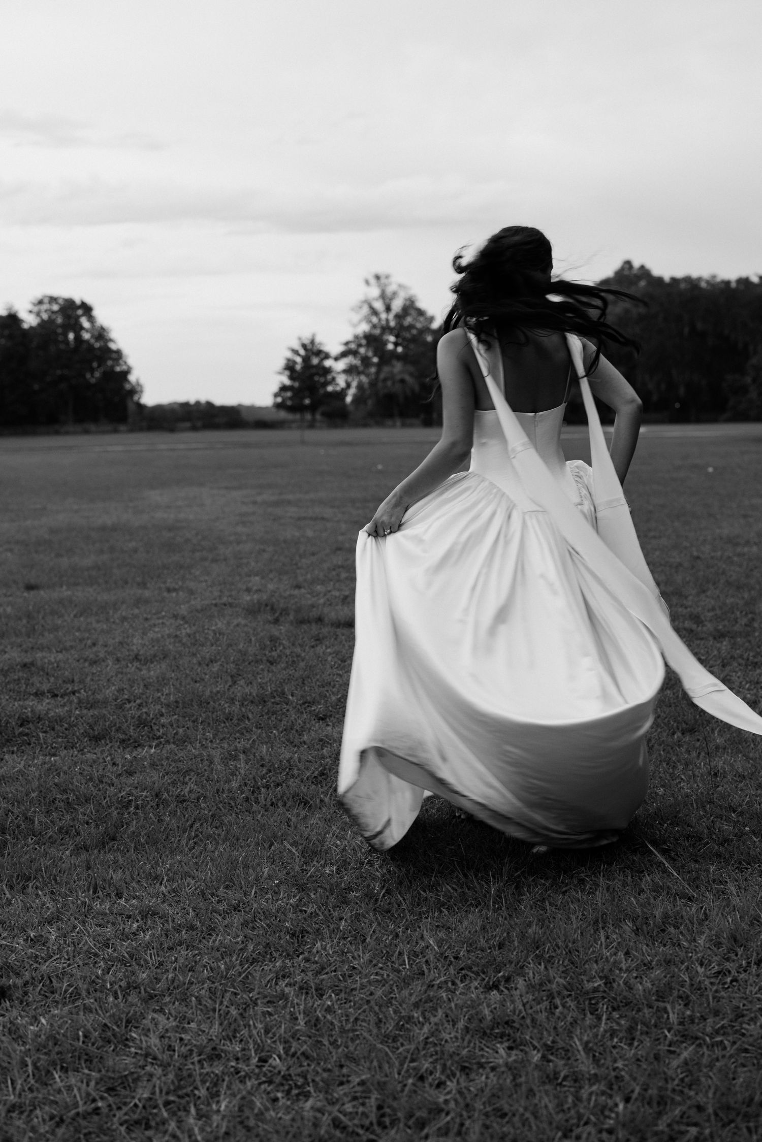 Black and white photo of bride running across open field in flowing white wedding gown.