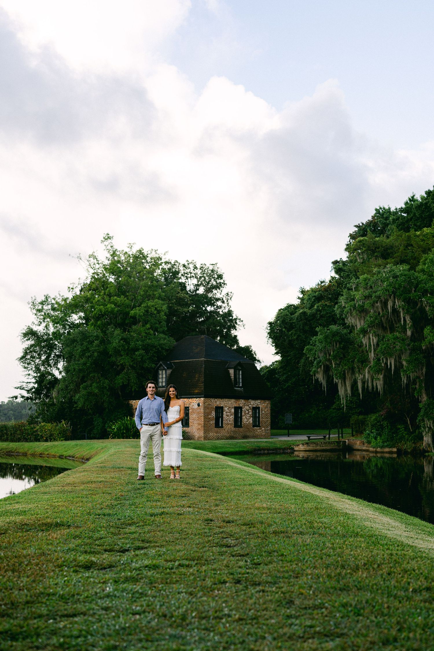 Couple standing on lush green lawn path between reflective ponds with historic brick building and Spanish moss trees behind them.