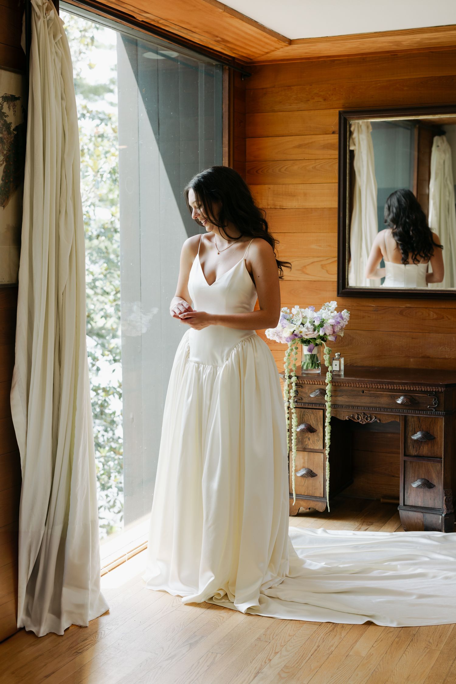 Bride in elegant ivory wedding gown standing by window with bouquet reflected in antique mirror.