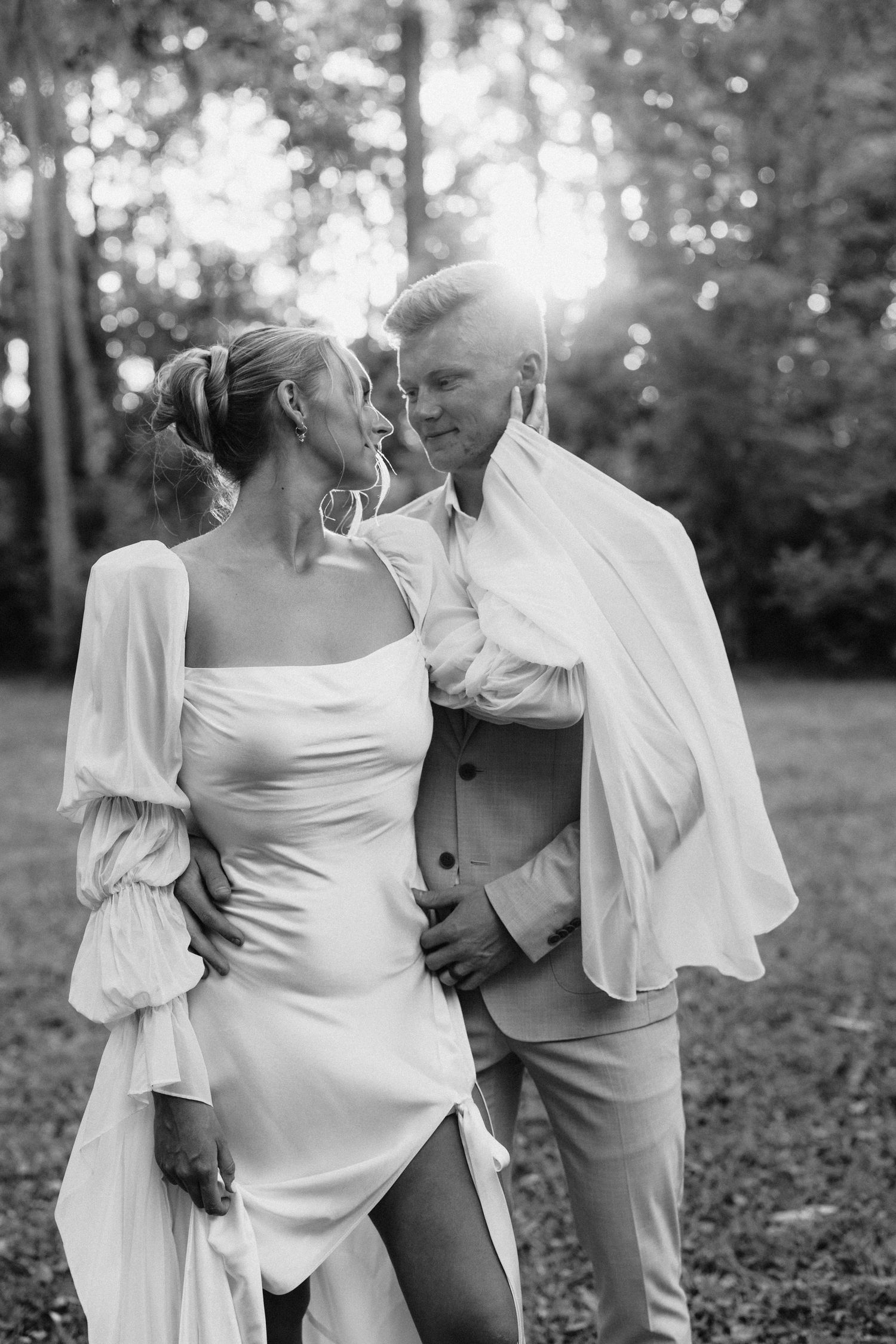 Black and white photo of bride and groom embracing in sunlit forest, bride in long-sleeve white gown.