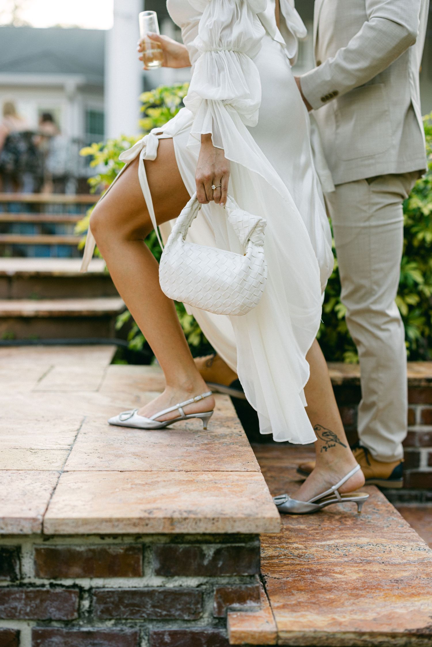 Bride in white dress with silver kitten heel shoes holding a woven clutch while climbing stone steps at wedding.