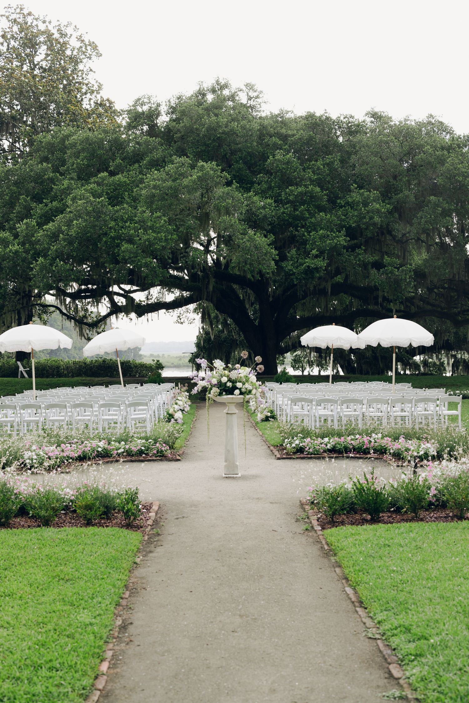 Elegant outdoor wedding ceremony setup with white chairs, floral arrangements, and large oak tree backdrop.