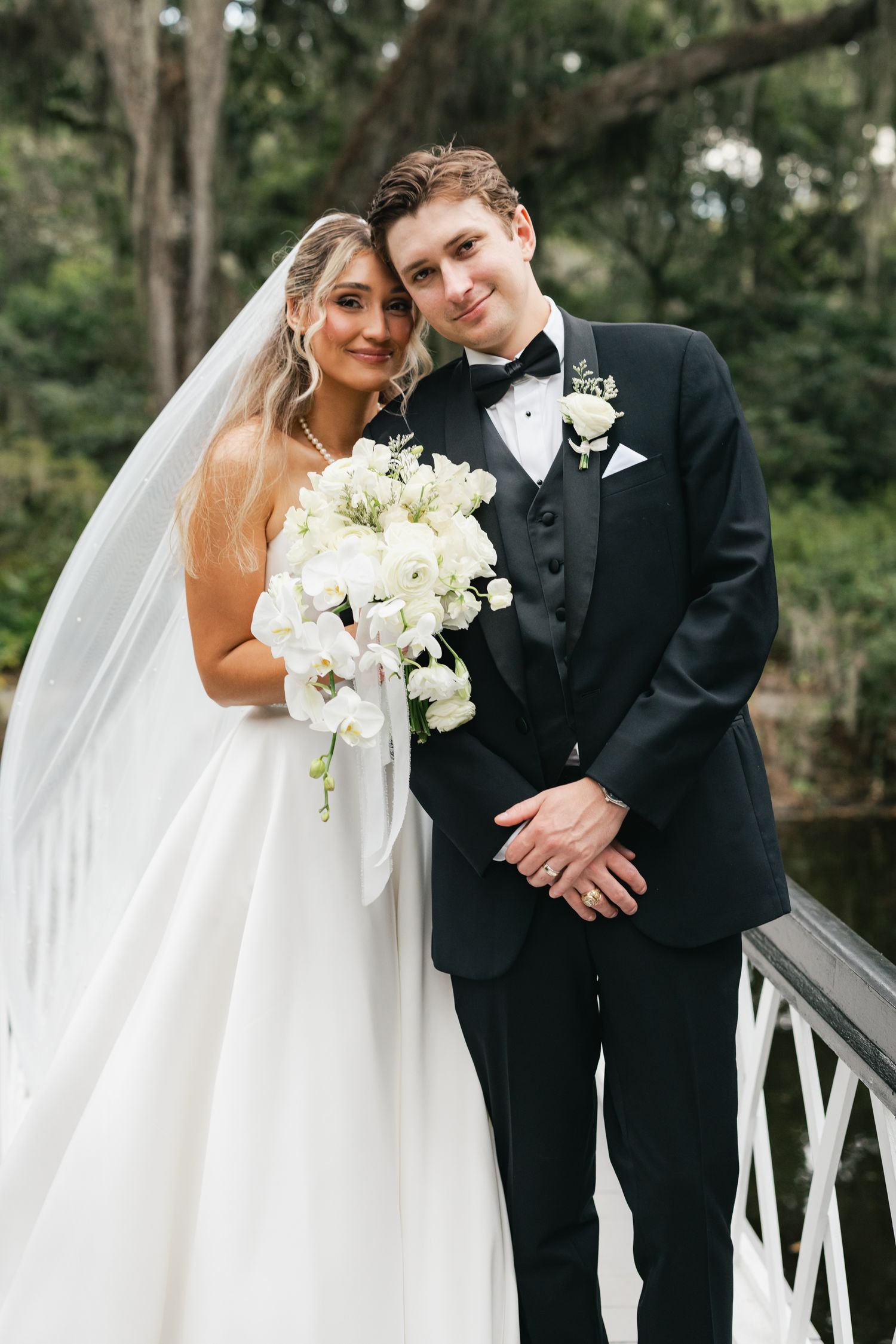 Bride in white gown with cascading bouquet and groom in black tuxedo posing on a bridge surrounded by lush greenery.