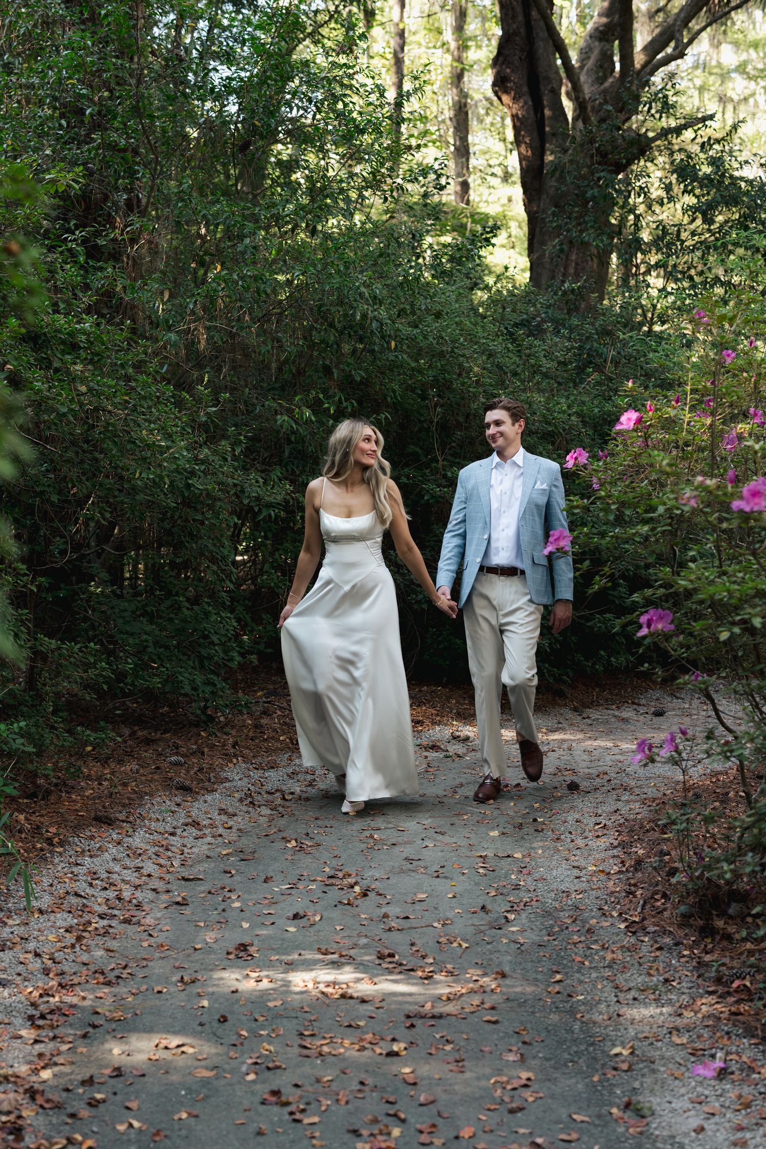 Bride and groom walk hand-in-hand along a wooded path surrounded by blooming azaleas.