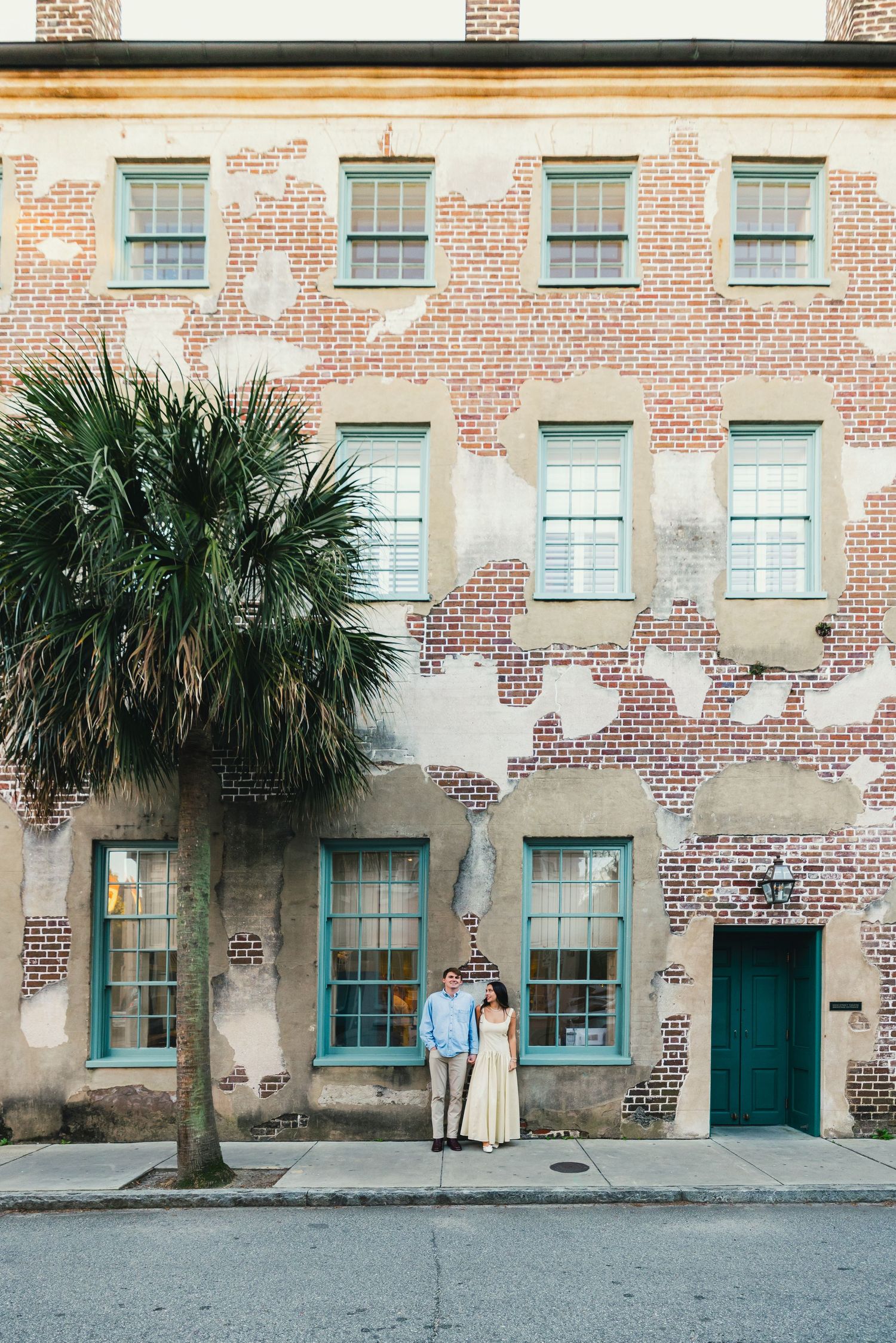 Couple poses in front of a historic Charleston brick building with turquoise windows and a palm tree.