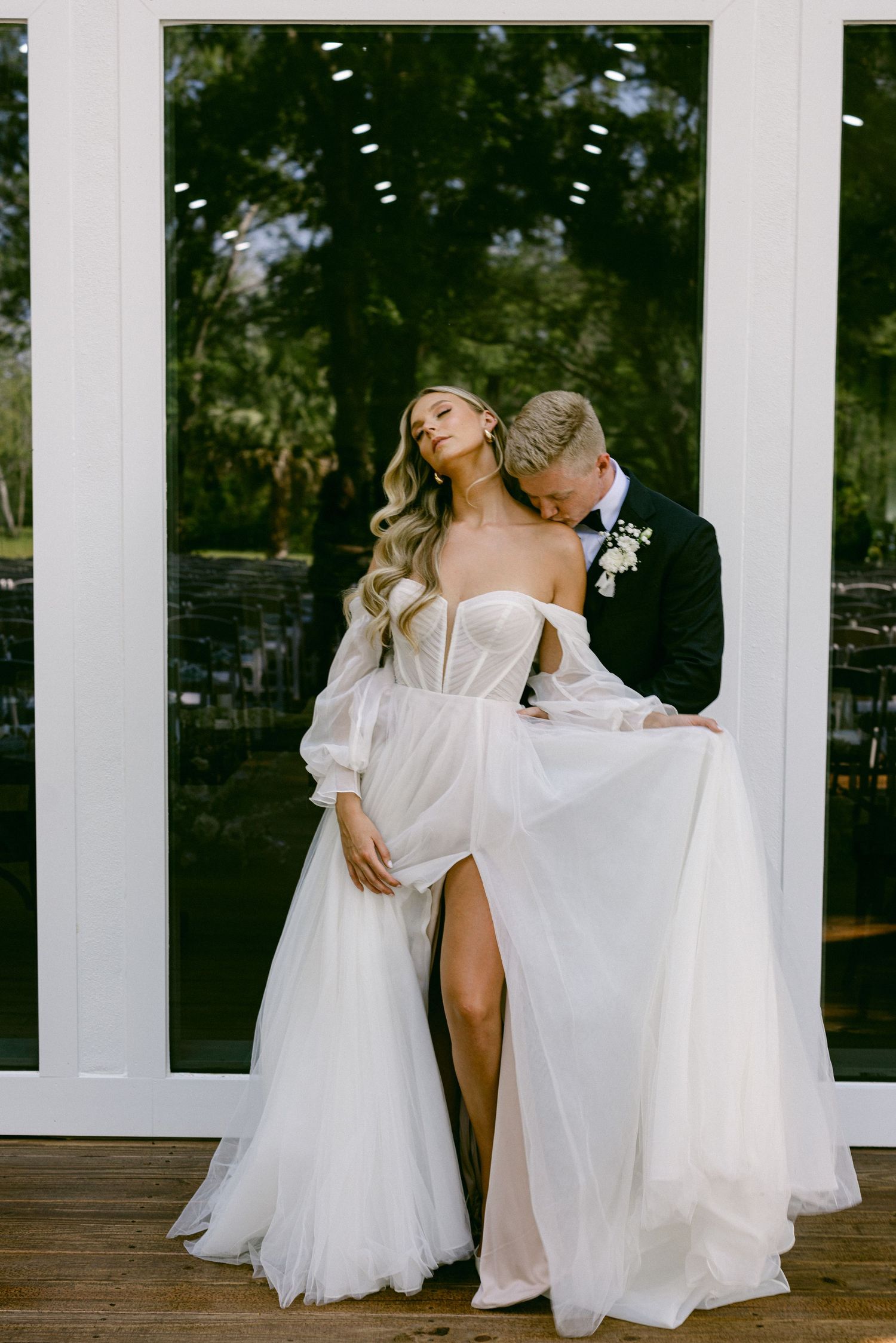 Bride in sheer white gown with slit embraced by groom in black suit before glass doors.