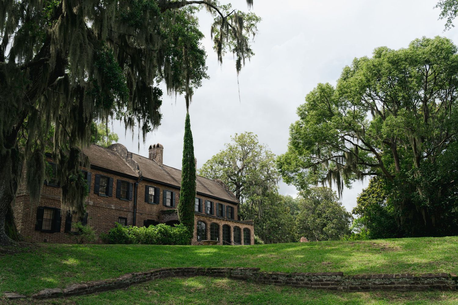 Historic brick plantation manor surrounded by ancient oak trees draped with Spanish moss on overcast day.