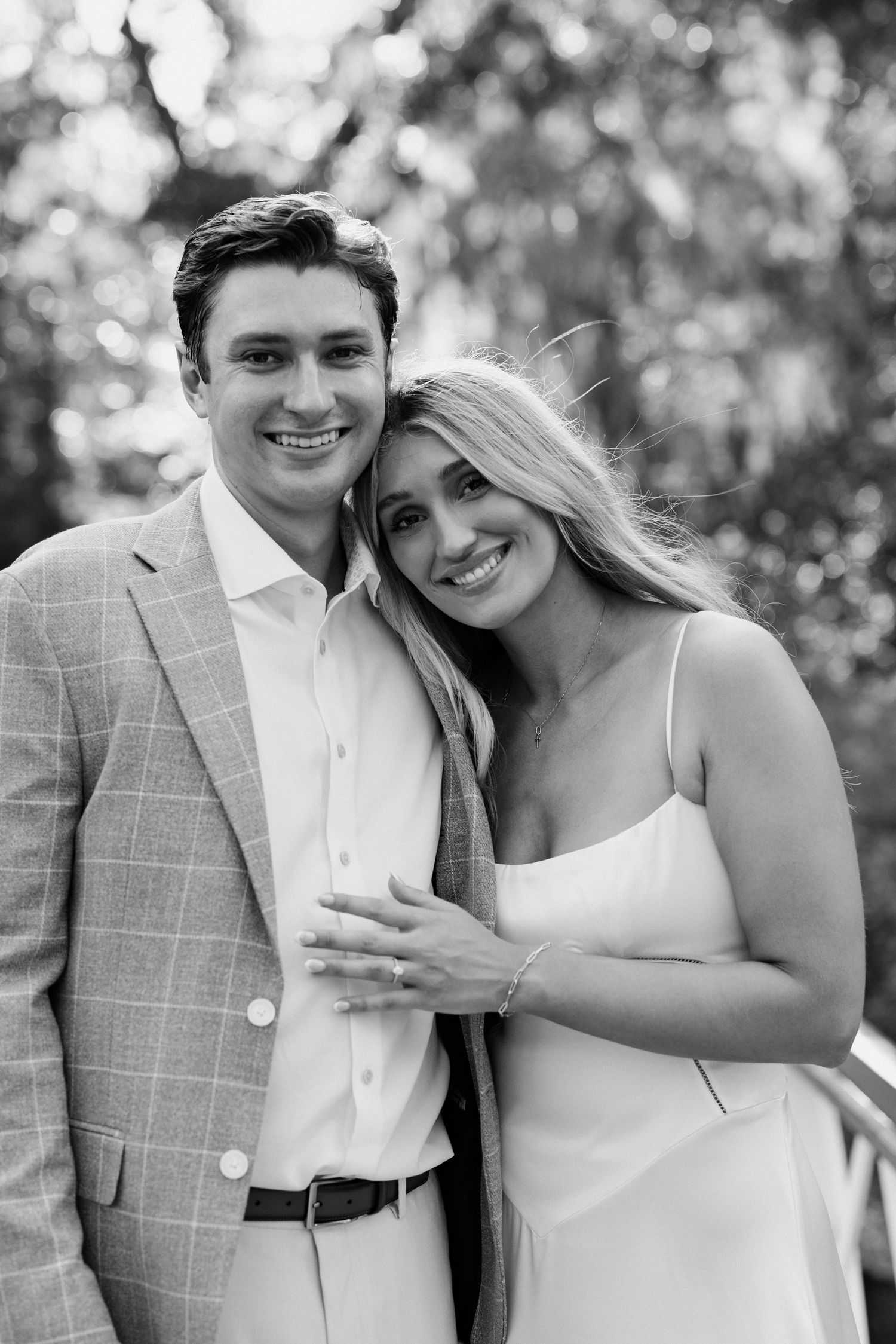 Black and white portrait of a smiling couple, man in plaid blazer and woman in white dress, posing outdoors.