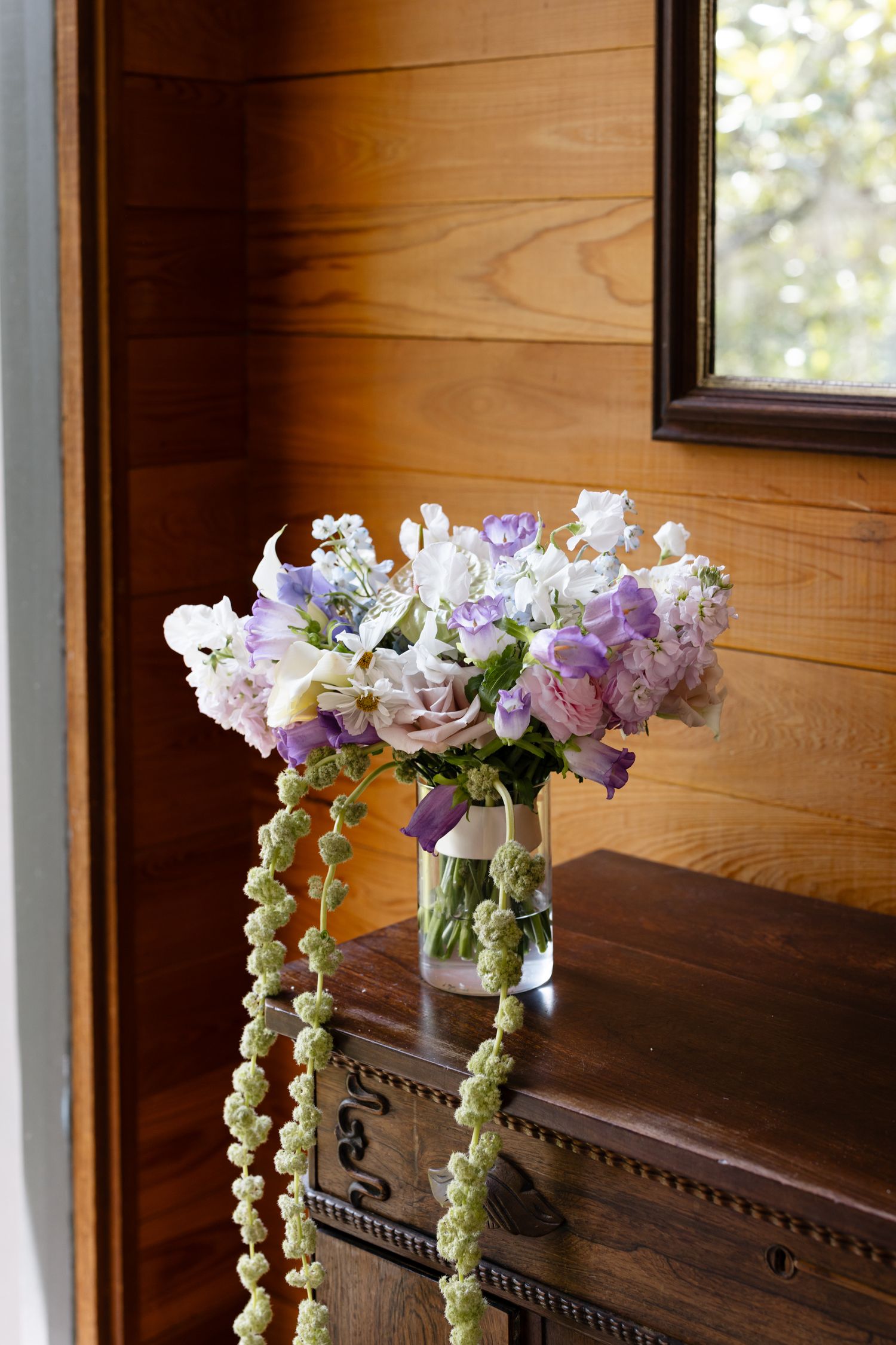 Elegant bridal bouquet of purple and white flowers in glass vase with cascading greenery on antique wooden dresser.