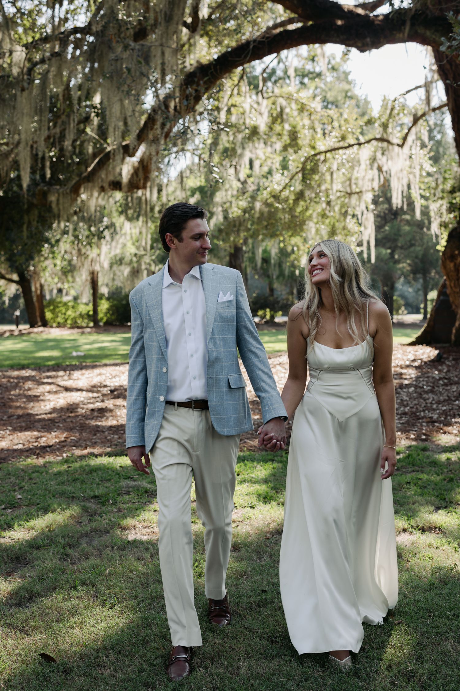 A couple walks hand-in-hand under Spanish moss-draped oak trees, the bride in a white gown and groom in a light blue suit.