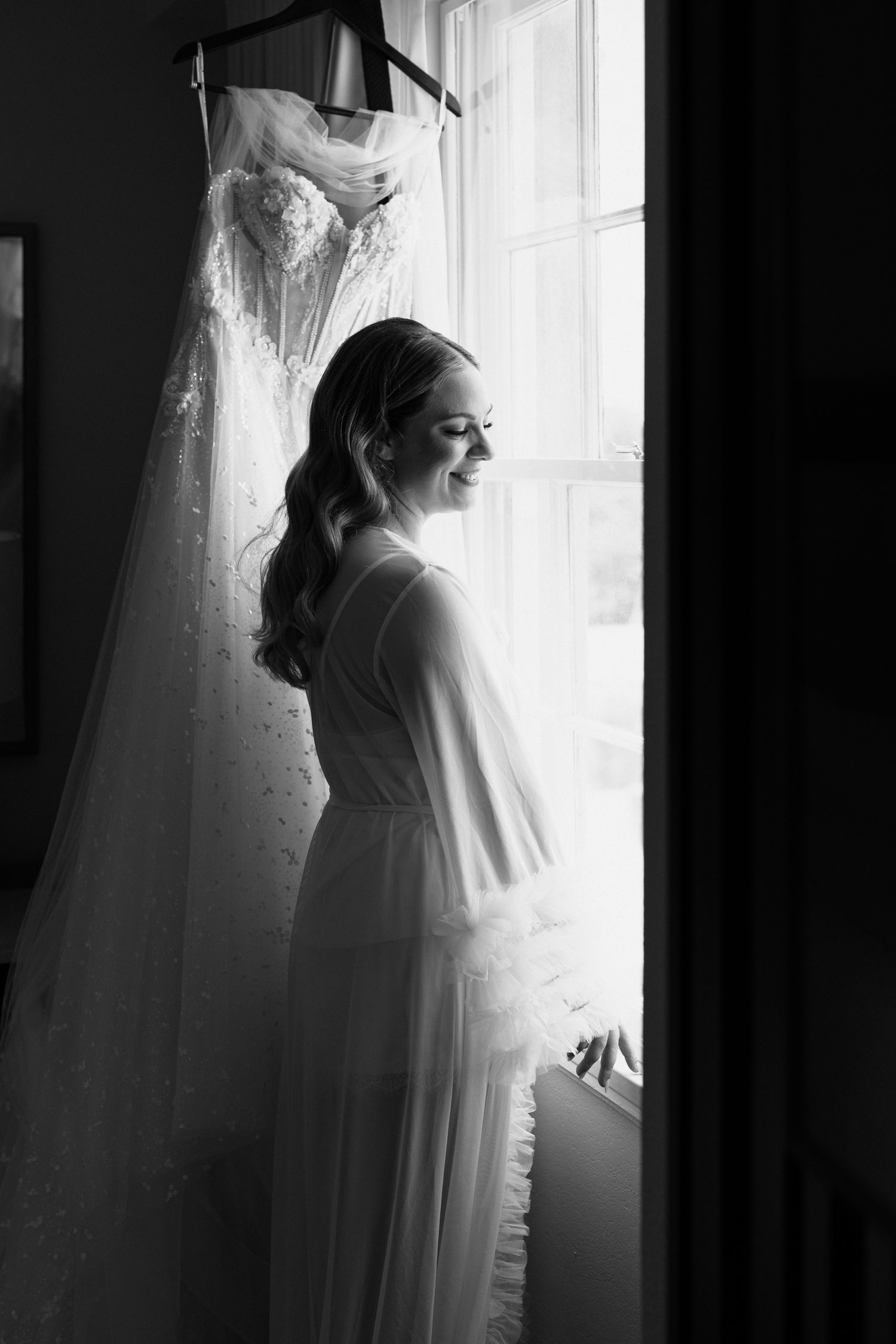 Black and white photo of a smiling bride in a flowing gown standing by a sunlit window with her wedding dress hanging nearby.