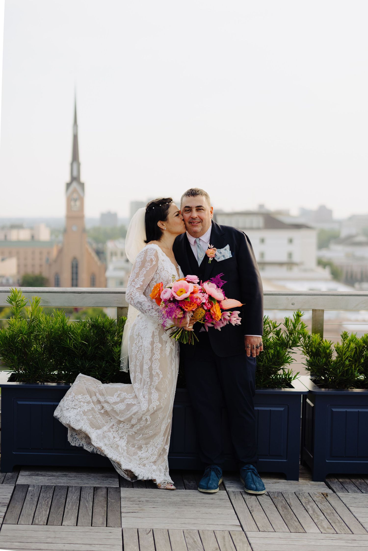 Bride kisses groom on rooftop with colorful bouquet, city skyline and church steeple in background.