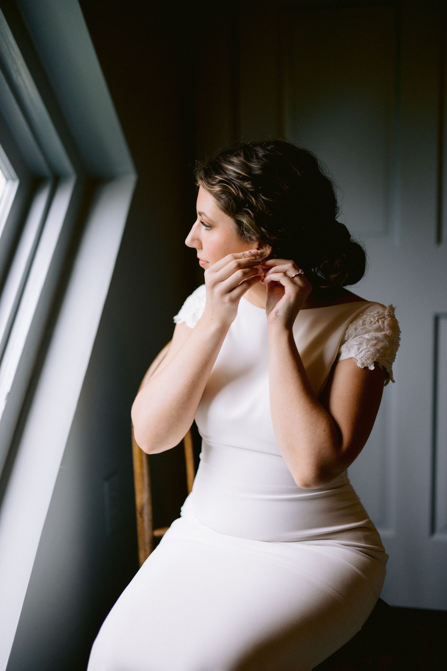 Bride in white fitted gown with lace sleeves putting on earrings by a window on her wedding day.