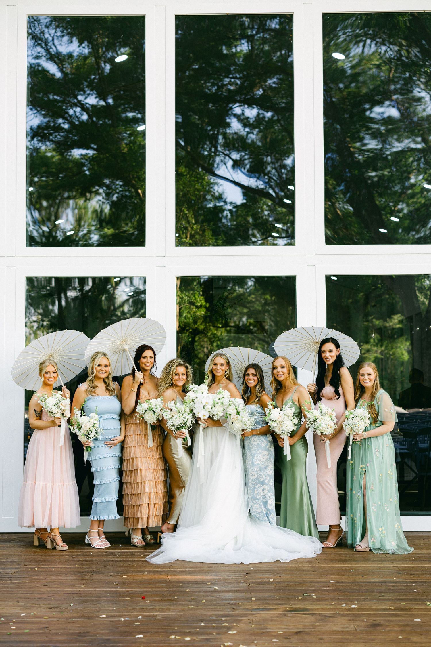 Bride with bridesmaids in pastel mismatched dresses holding white bouquets and parasols outside a venue.