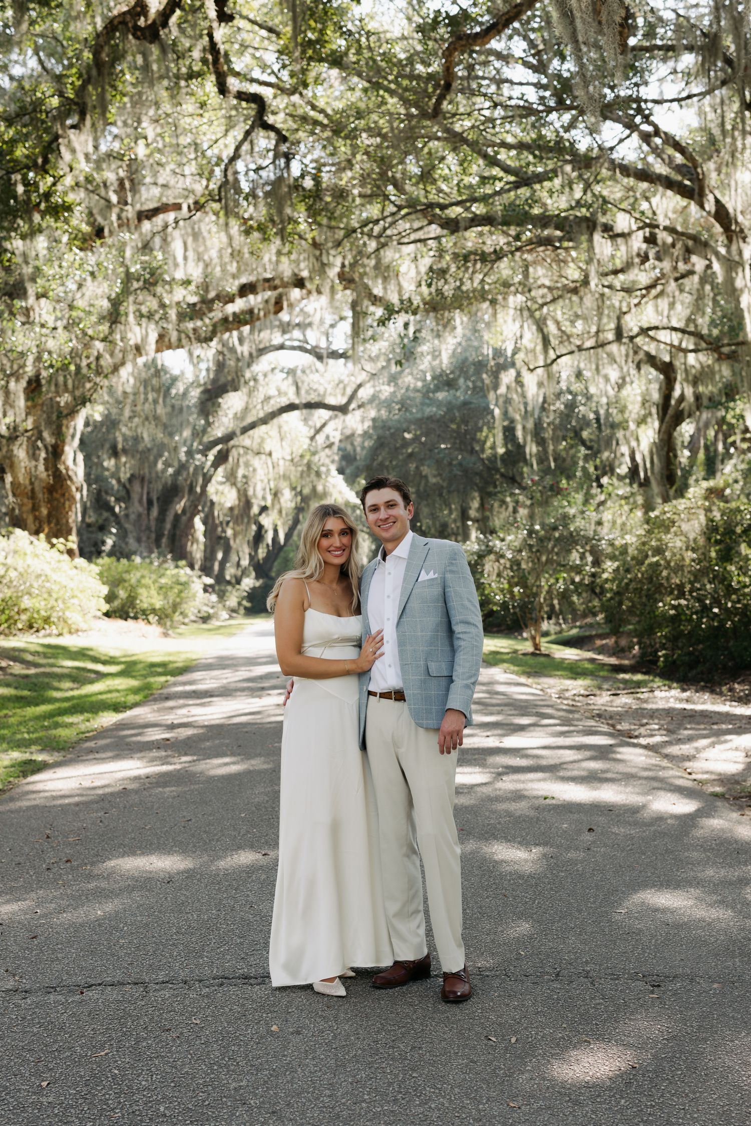 Couple in wedding attire pose on sunlit path beneath Spanish moss-draped oak trees.