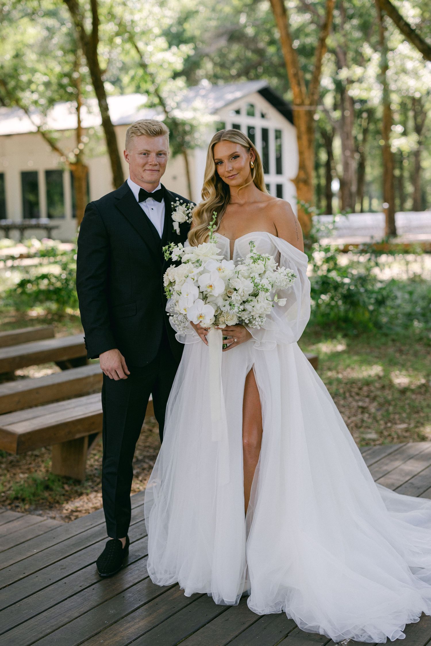 Bride in off-shoulder tulle gown with slit holds white bouquet beside groom in black tuxedo outdoors.