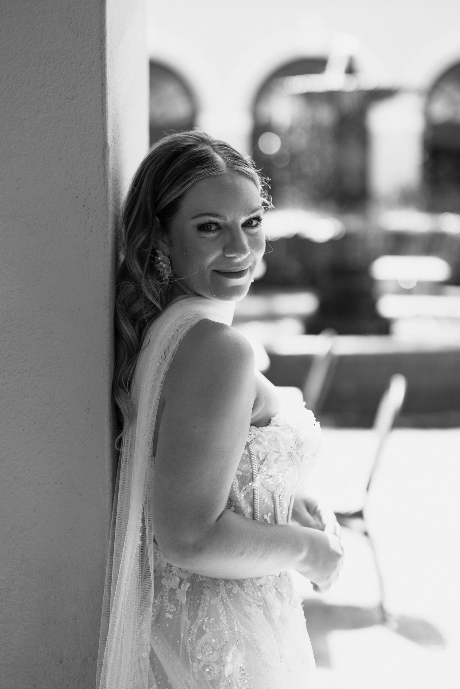 Black and white portrait of a smiling bride in lace gown and veil leaning against a wall outdoors.