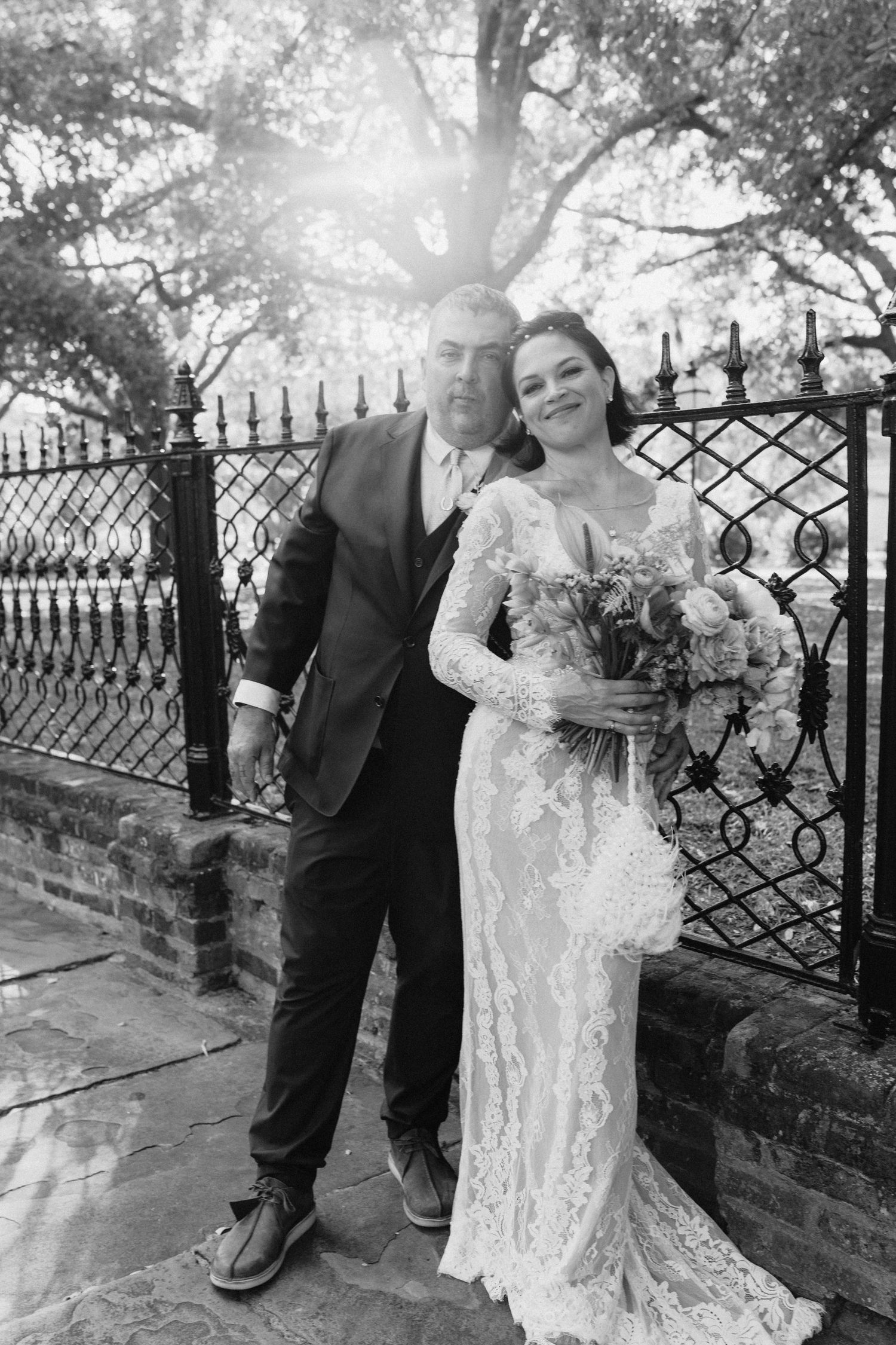 Black and white photo of a bride in lace gown holding bouquet posing with groom in suit by ornate iron fence.