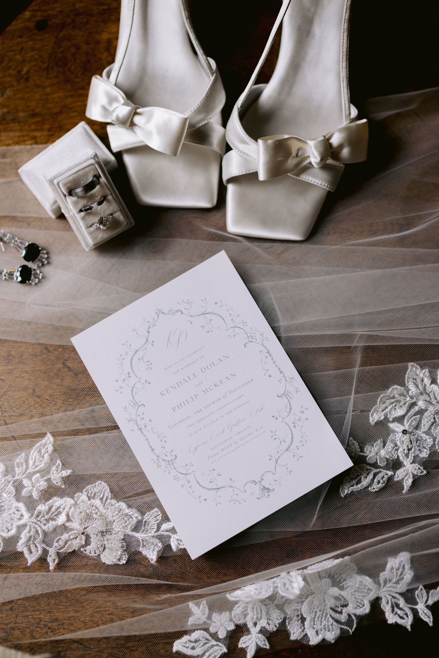 Elegant bridal flat lay featuring ivory bow heels, lace veil, wedding invitation, and crystal earrings on wood.