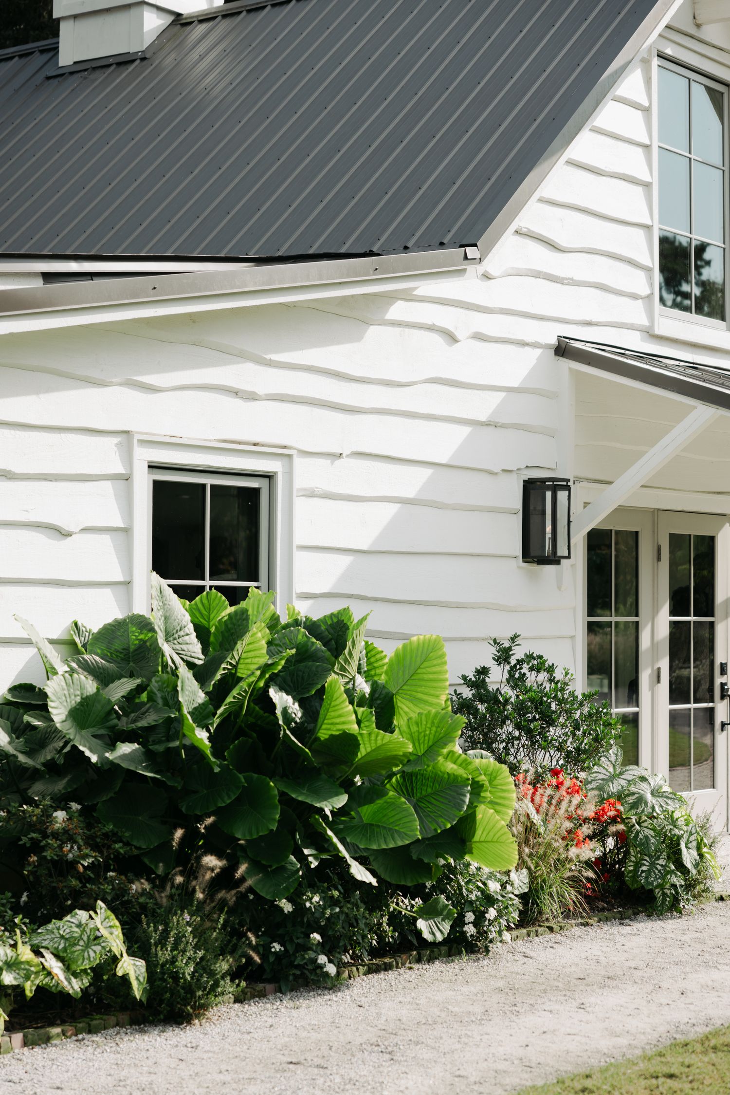 White farmhouse-style wedding venue exterior with metal roof, lush tropical plants and French doors.