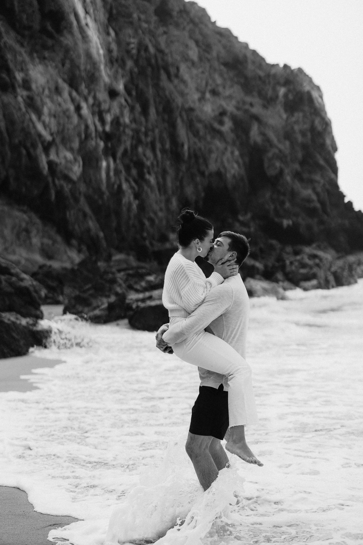 Black and white photo of man lifting woman in ocean waves near rocky cliff.