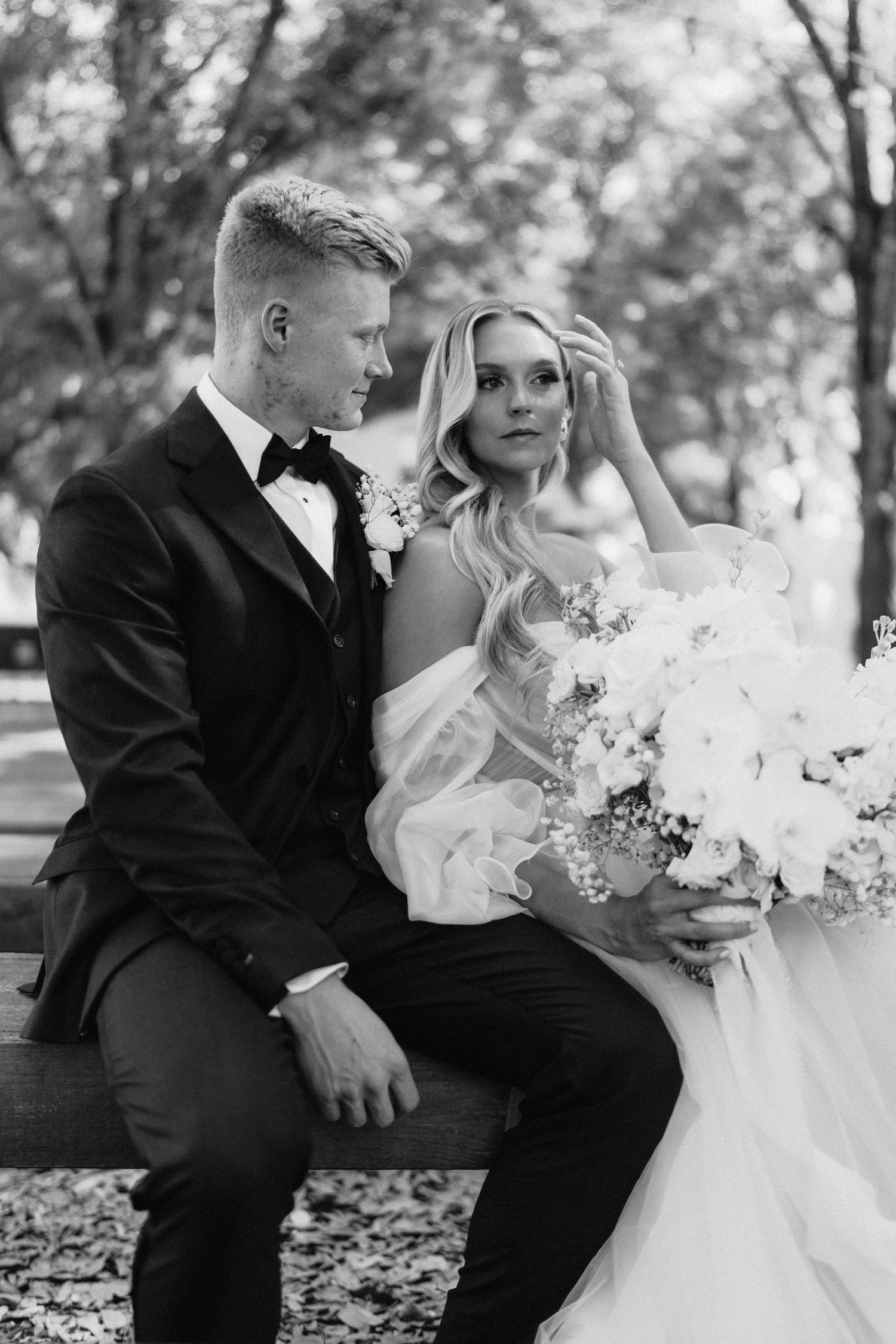 Black and white portrait of bride and groom seated on bench, bride holding large white bouquet.