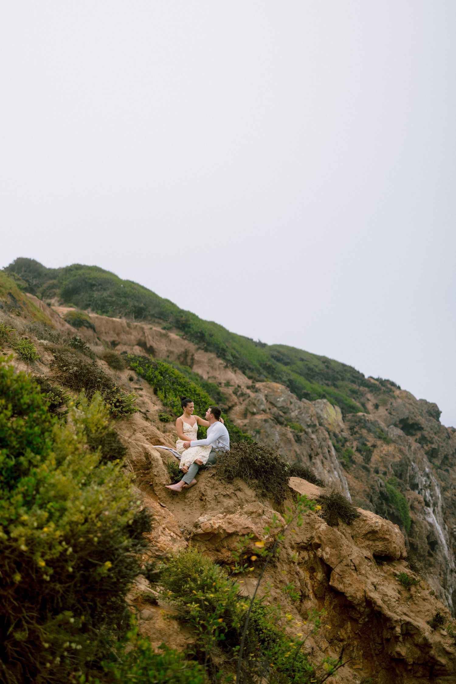 Couple sitting together on rocky hillside cliff surrounded by lush green coastal vegetation.