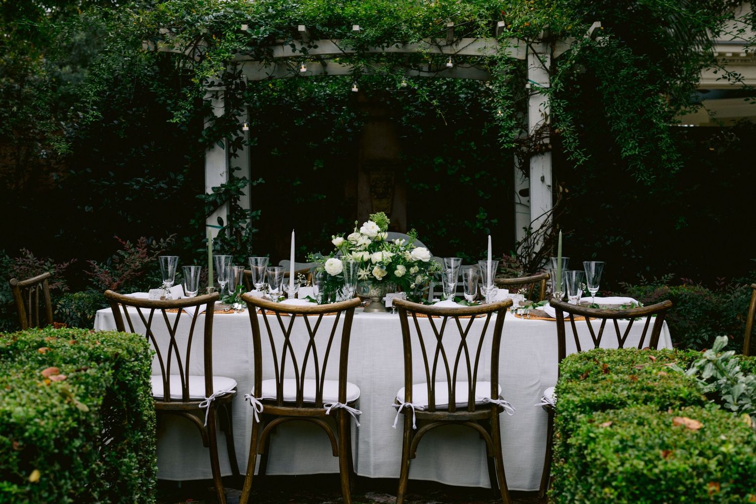 Garden wedding reception table set with white linens, floral centerpiece, candles, and wooden chairs amid lush greenery.