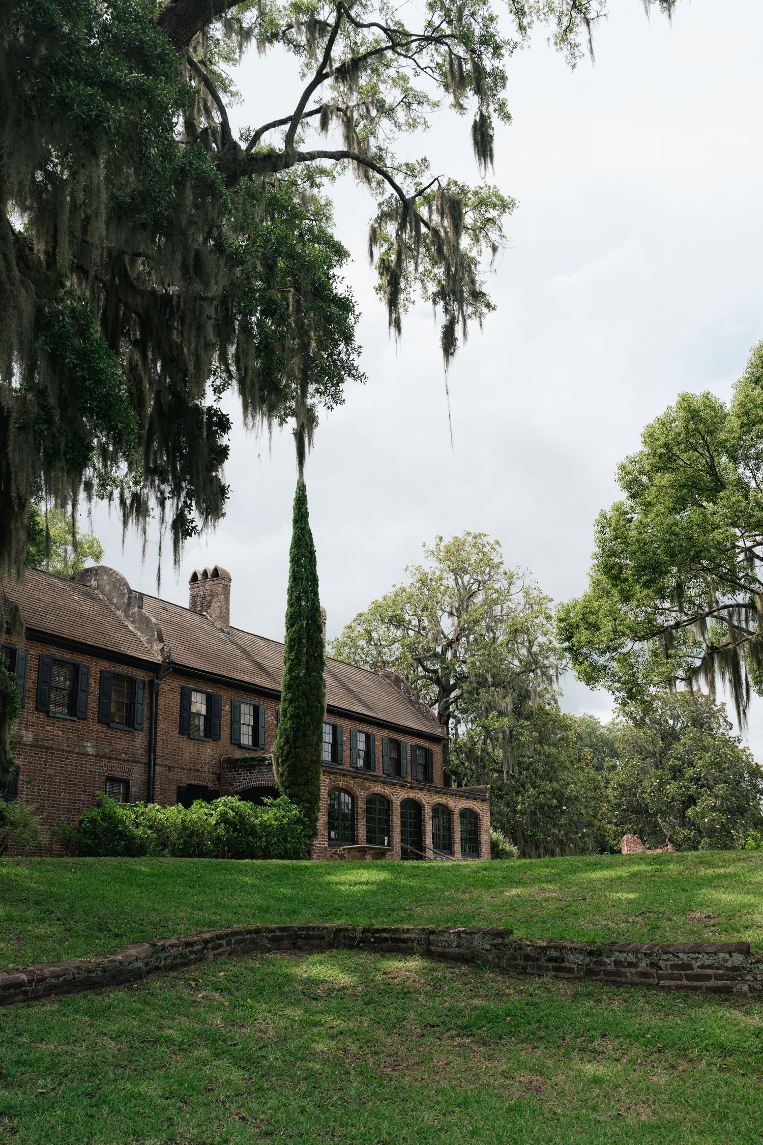 Historic brick plantation house with Spanish moss-draped oak trees and lush green lawn in the American South.