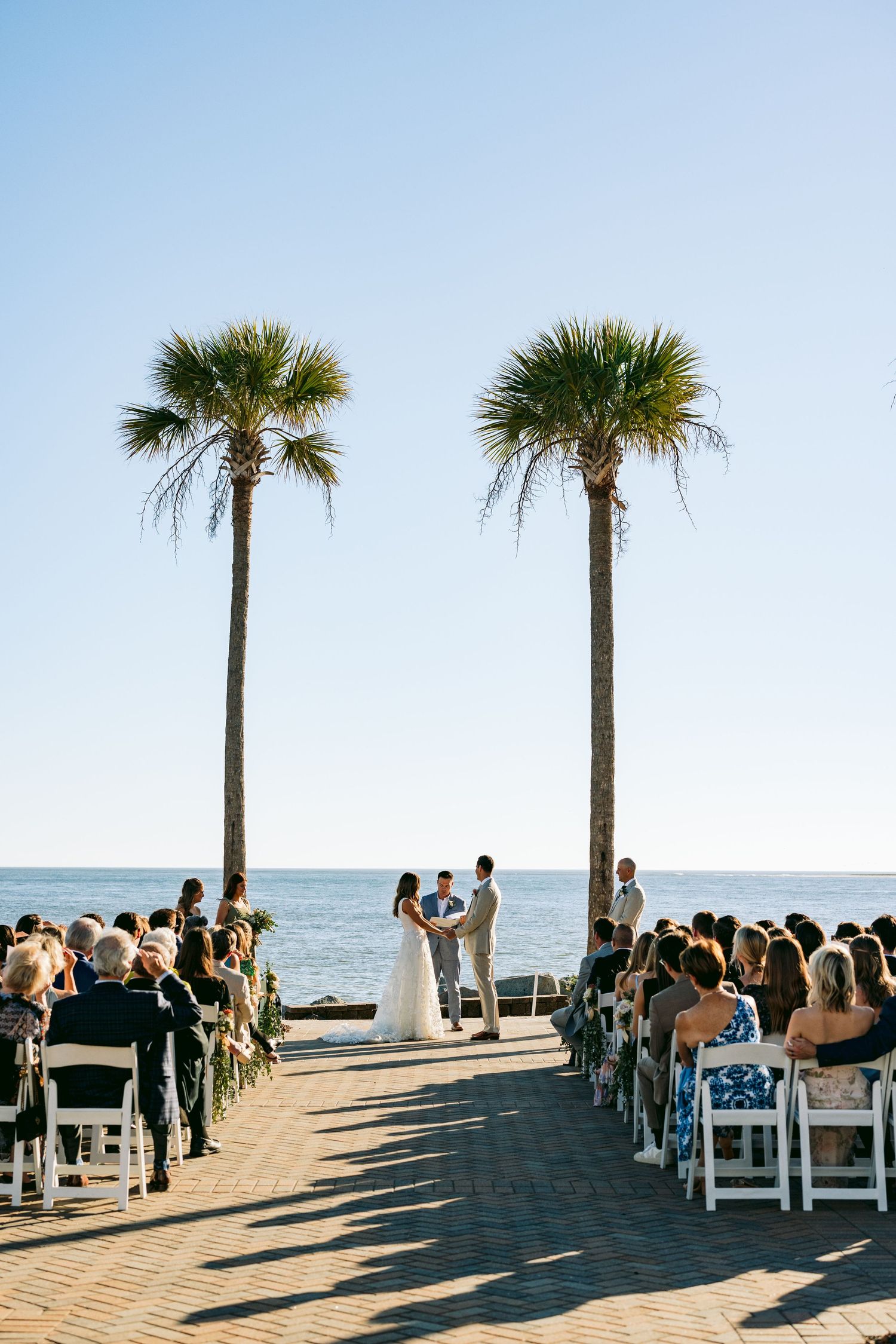 Outdoor beach wedding ceremony with couple at altar between two tall palm trees, guests seated in white chairs by the ocean.