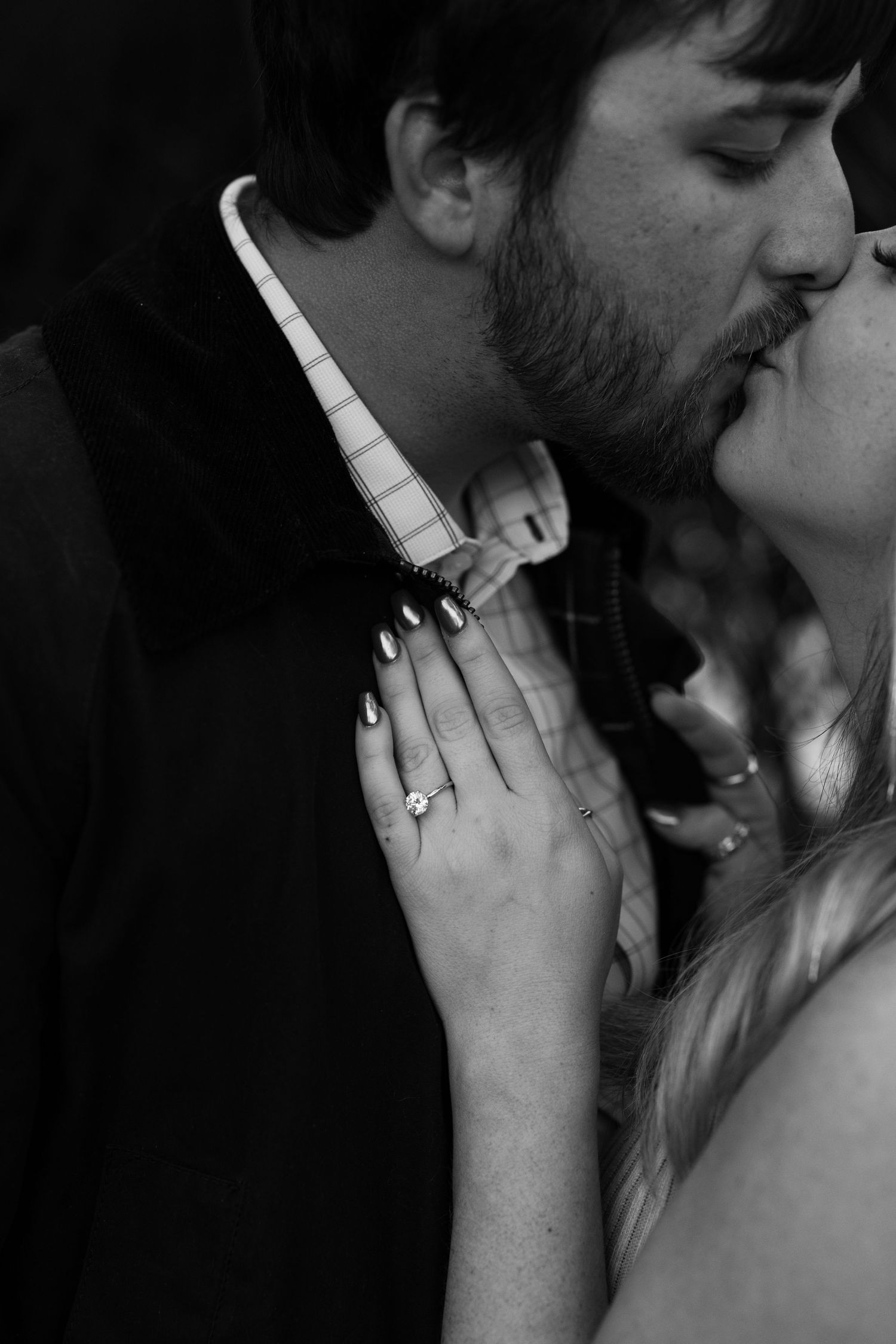 Black and white photo of a couple sharing a romantic kiss, woman showing engagement ring with dark painted nails.