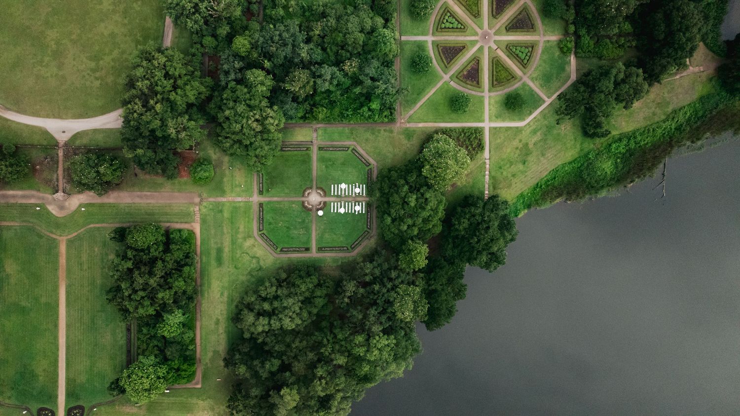 Aerial view of formal garden with circular patterns beside a tranquil river surrounded by green trees.