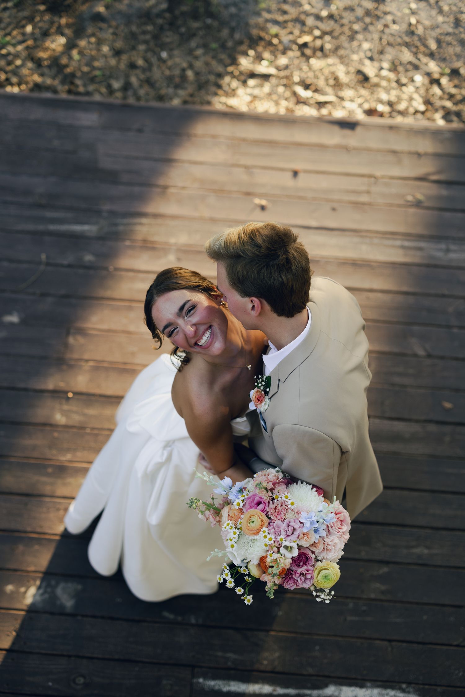 Bride smiling up at groom as he kisses her cheek, holding a colorful bouquet, photographed from above on wooden deck.