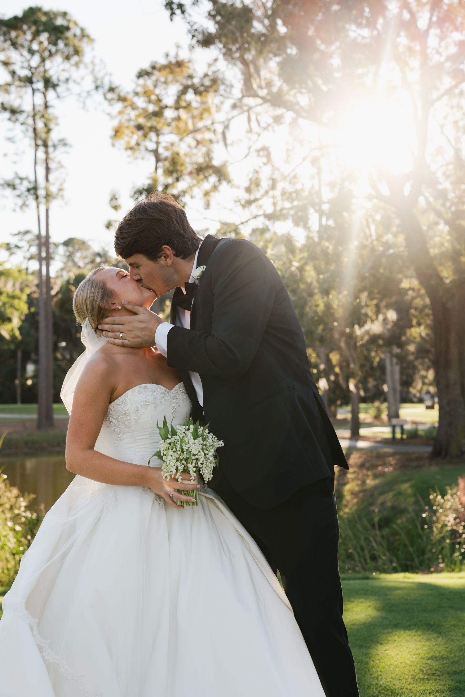 Bride and groom share a romantic kiss outdoors, with sunlight streaming through trees, bride holding a white bouquet in a strapless gown.
