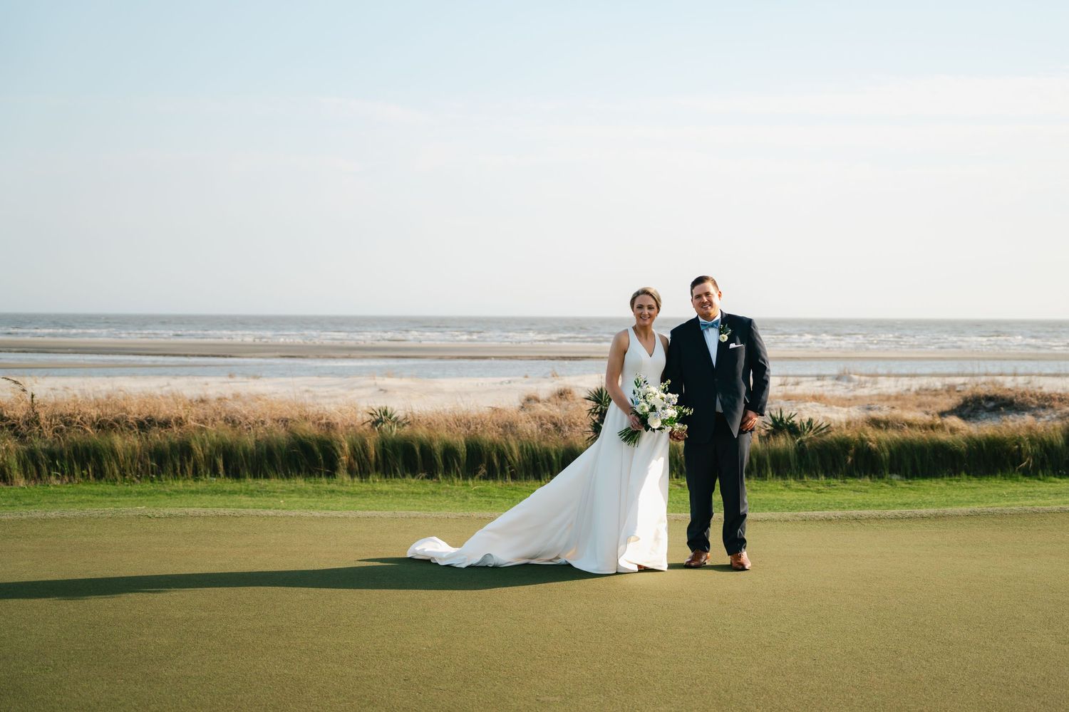 Bride and groom posing on golf course green with scenic ocean beach backdrop on wedding day.