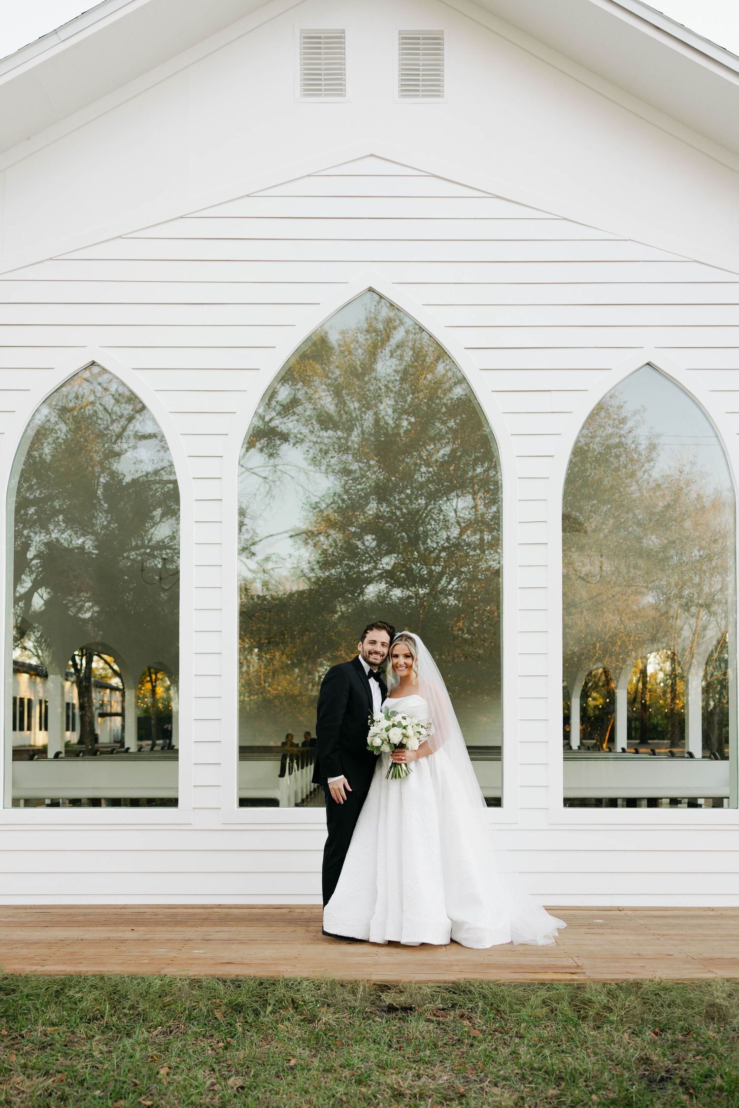 Bride and groom pose in front of a white chapel with Gothic arched windows on their wedding day.